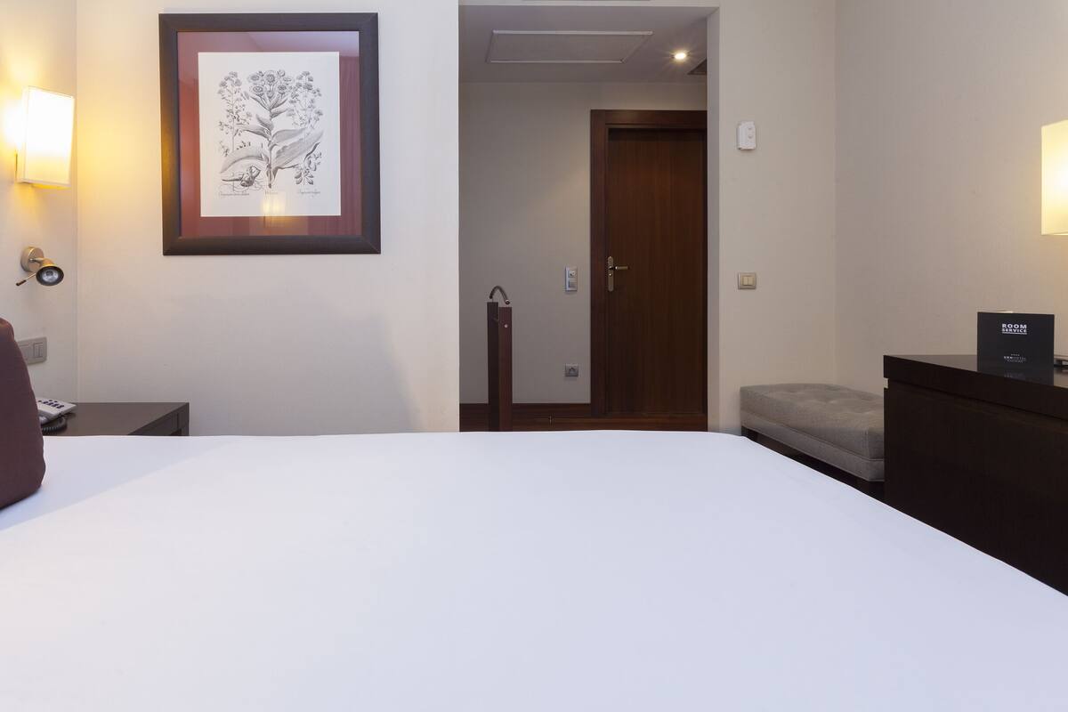 Ein Hotelzimmer mit einem Bett, einem Nachttisch, einer gerahmten Pflanzenillustration, einer Tür und einem kleinen Tisch mit einer Lampe.