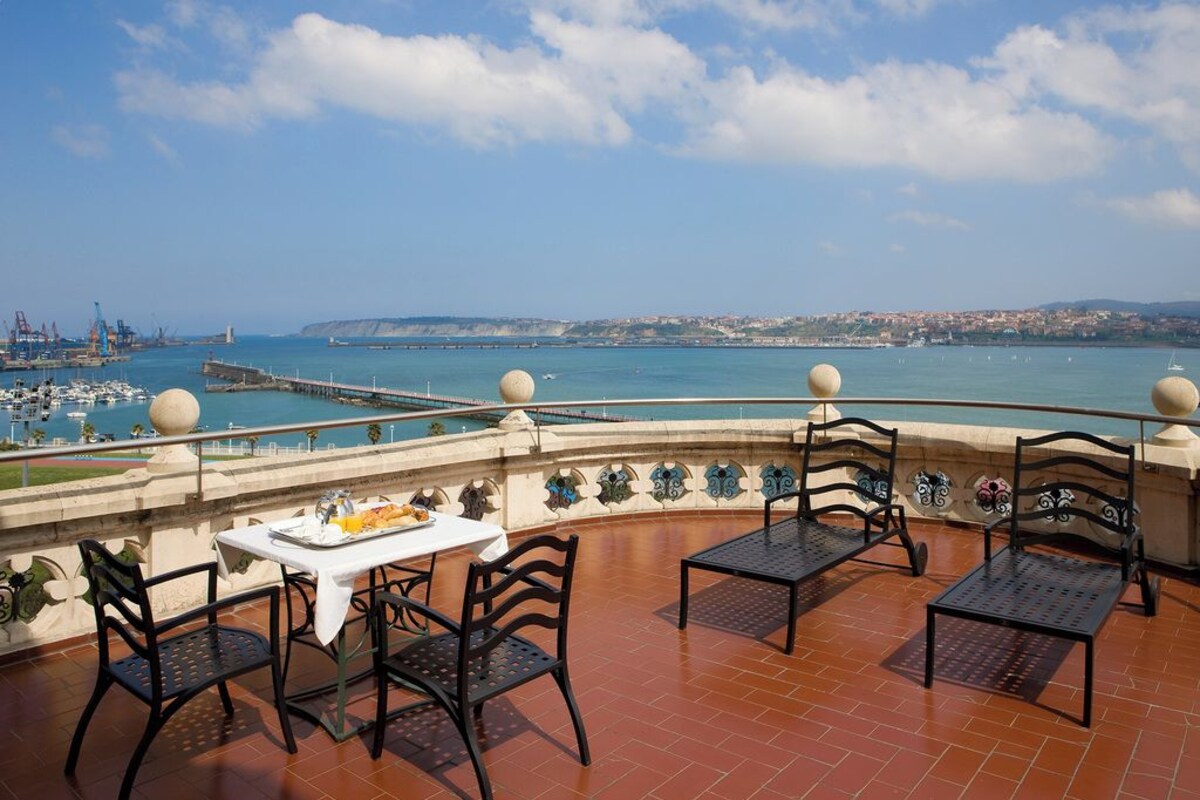 A terrace with a table and chairs, overlooking a coastal area with a pier and boats.
