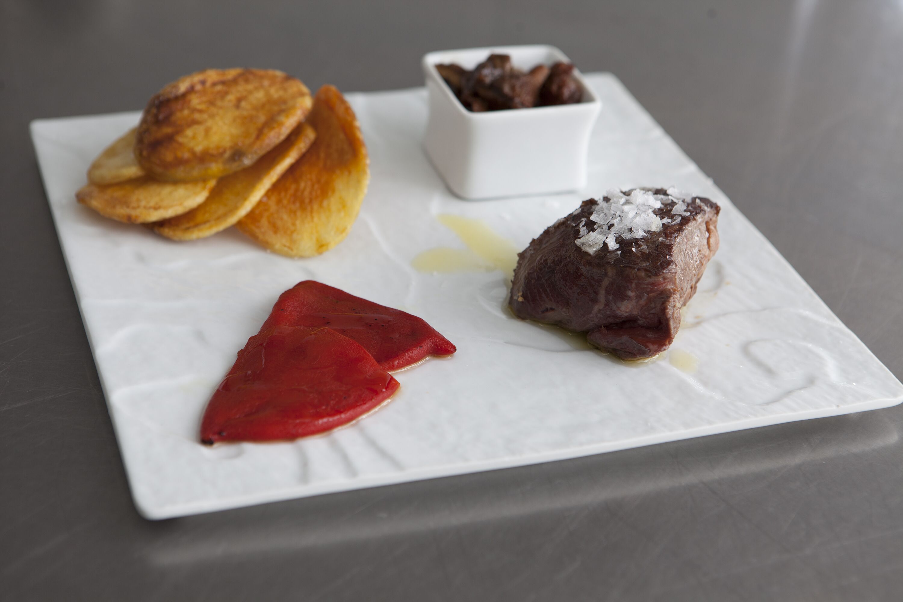 A white plate with food items including a piece of meat, a red item, and a small bowl of chocolate.