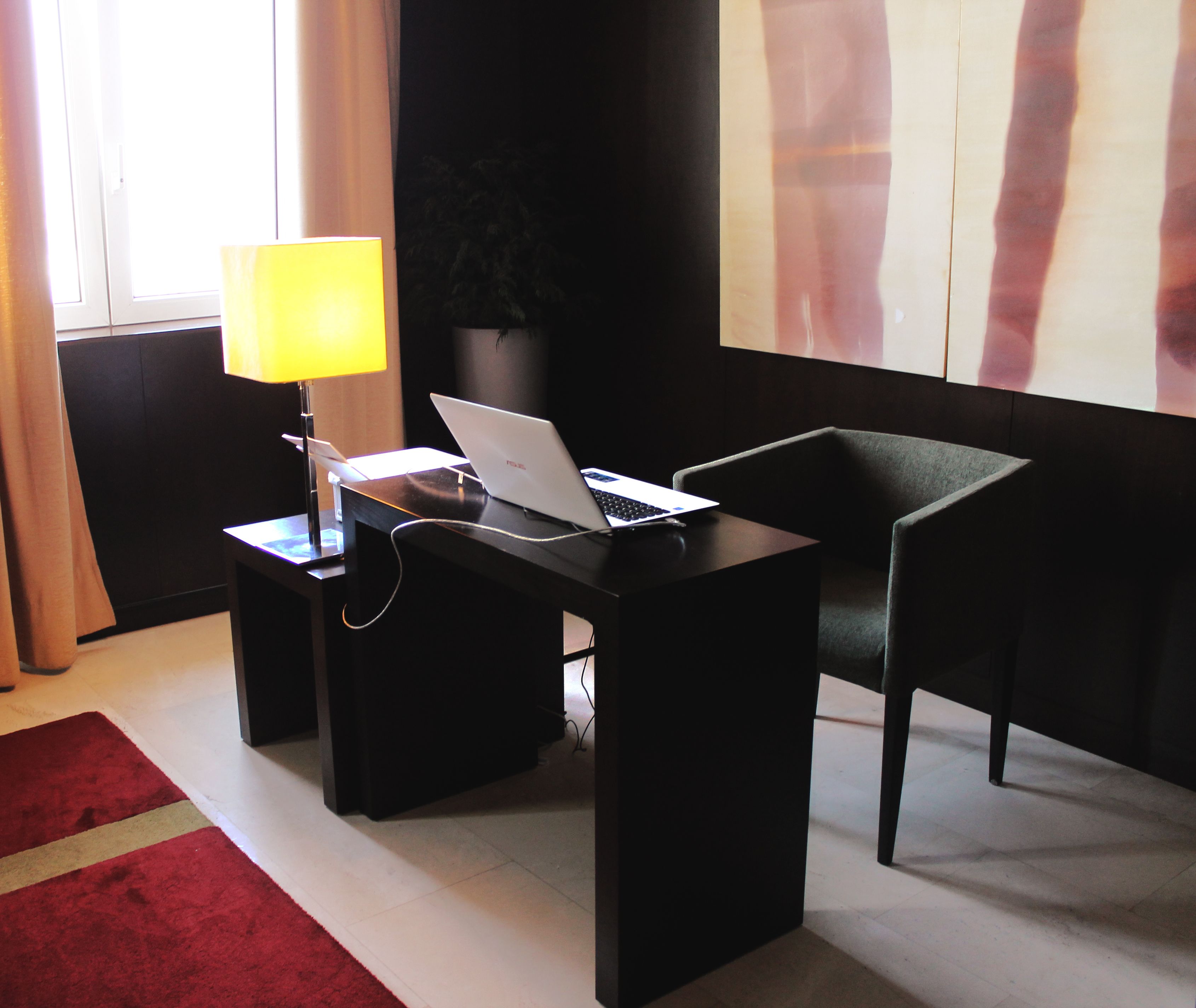 A modern hotel room with a desk, laptop, lamp, and a red rug.