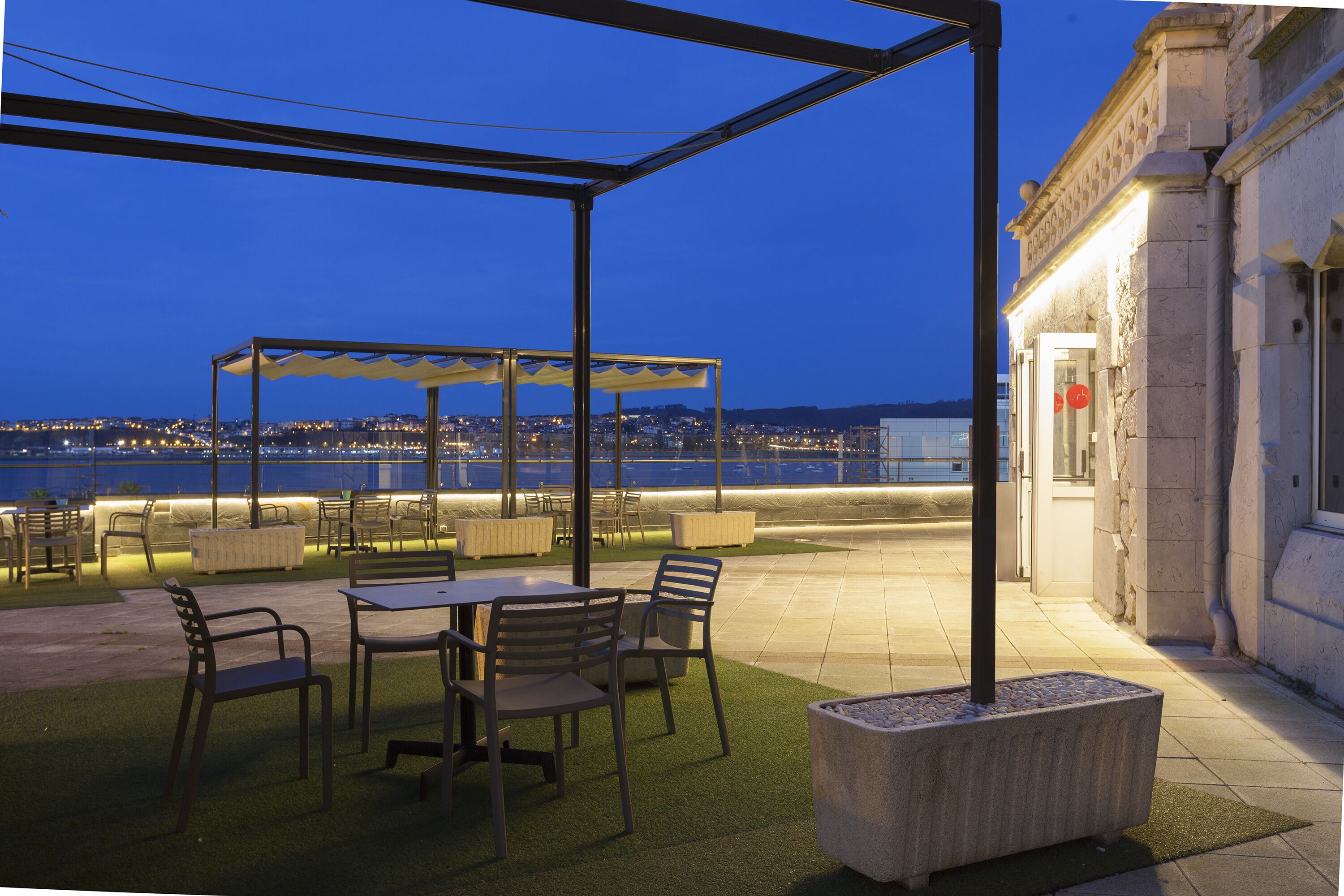 Een terras met tafels en stoelen, een pergola, en uitzicht op de stad en het water in de schemering.