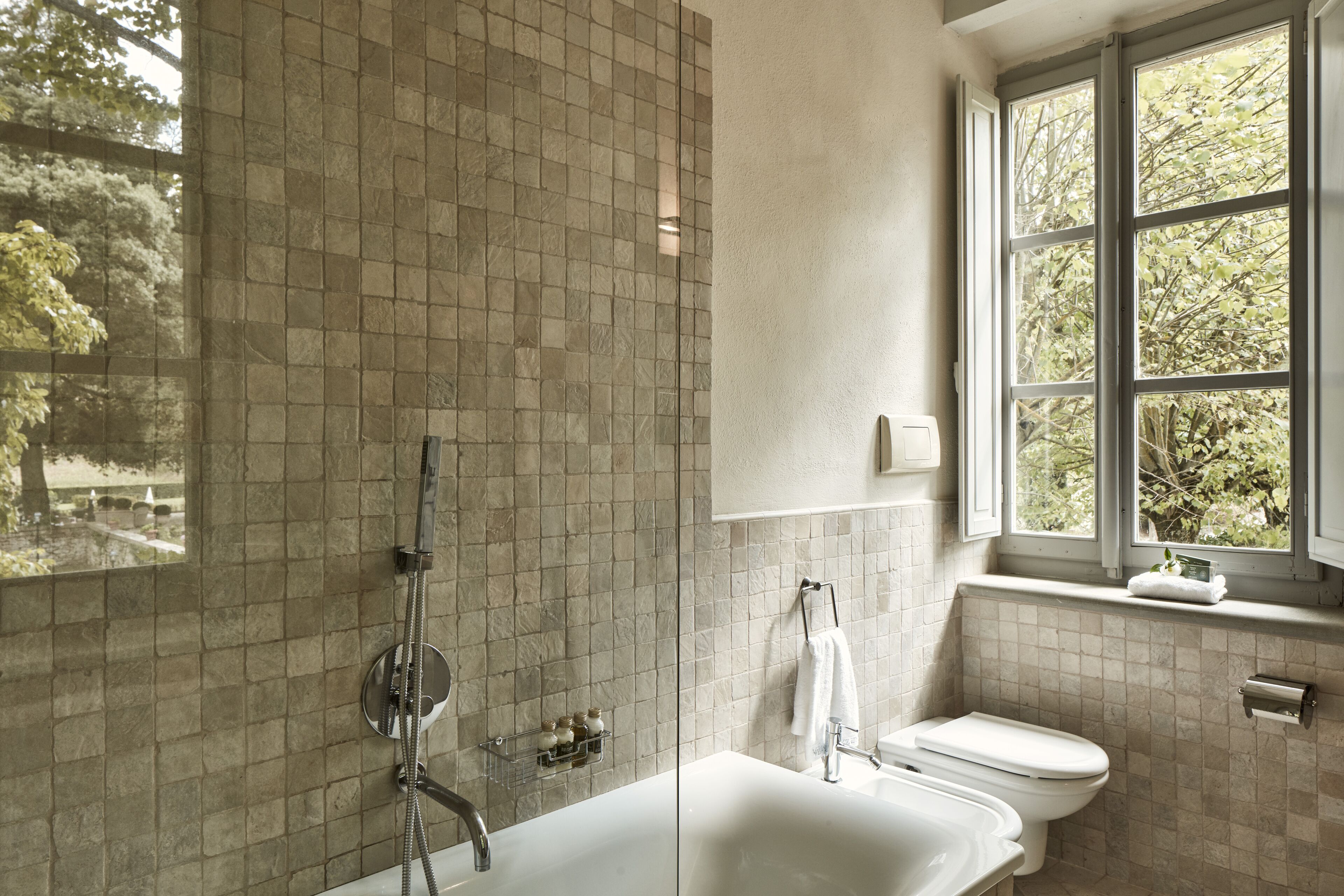 Un baño con bañera, inodoro y ventana con vista a árboles.