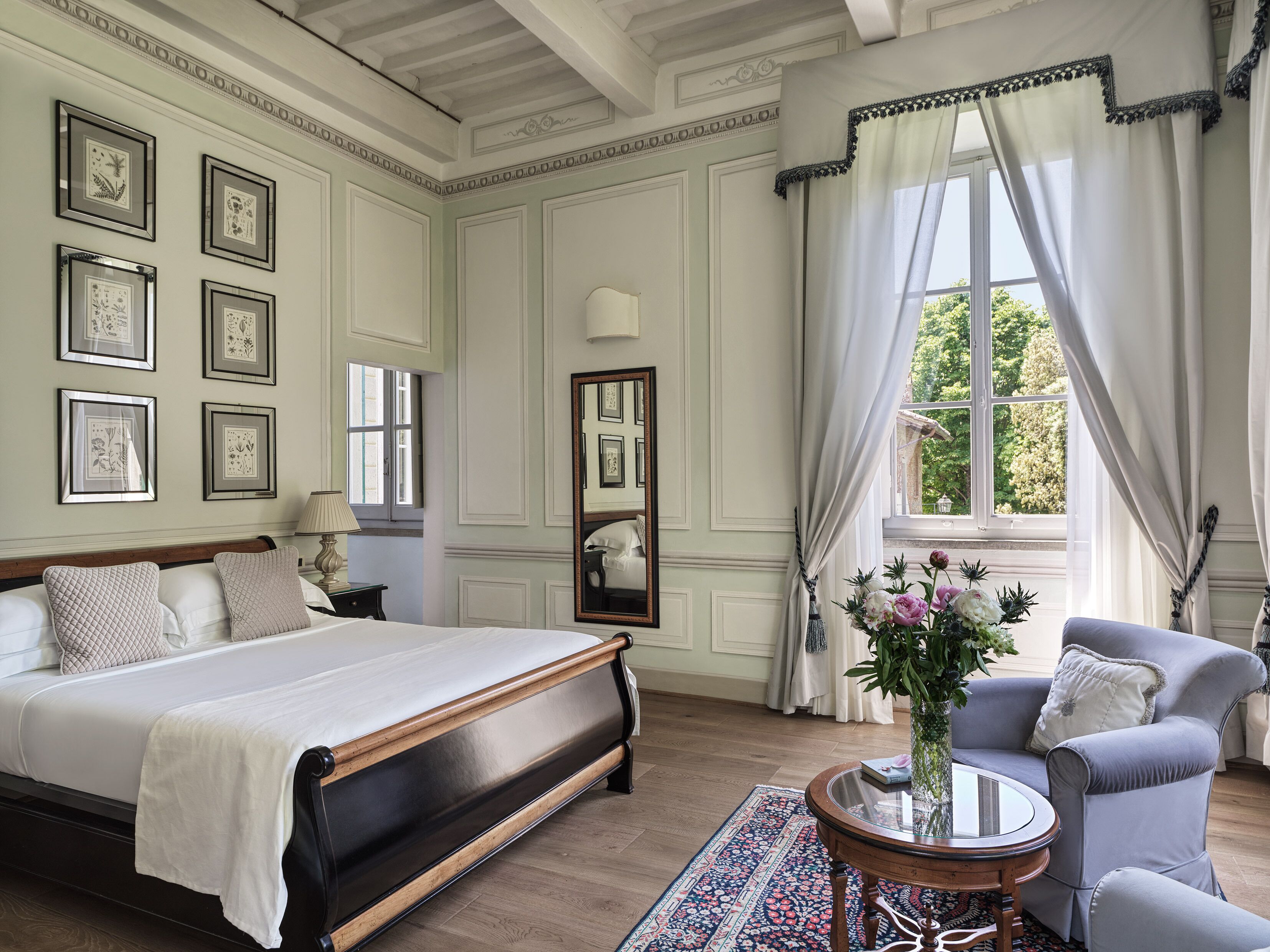 A spacious bedroom with a large bed, a sitting area with a chair and table, and a window with curtains.