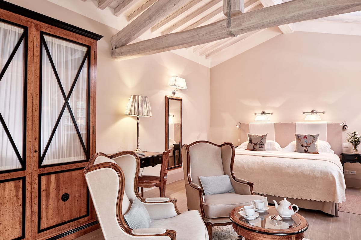 A bedroom with a large bed, two armchairs, a wooden wardrobe, a desk, and a bedside table with a lamp.
