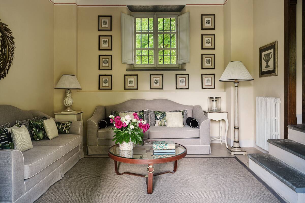 A living room with a grey sofa, a coffee table with a vase of flowers, framed pictures on the wall, and a window with a view of greenery.