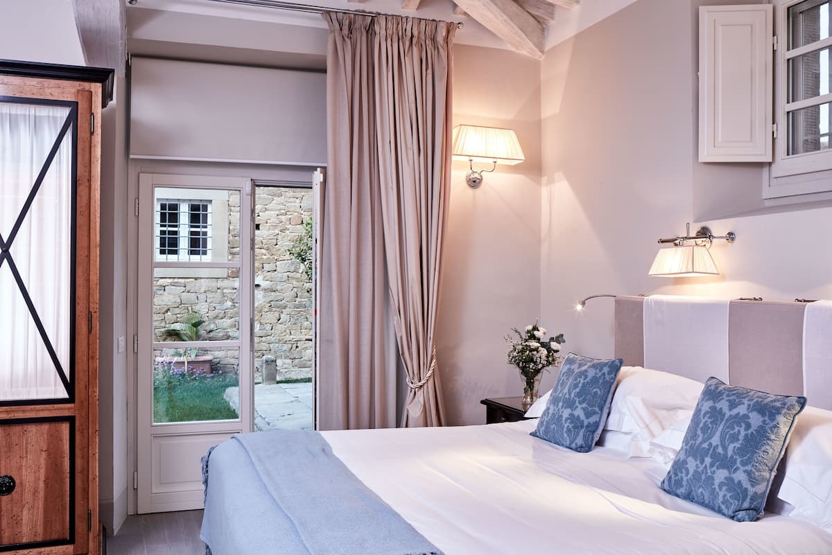 A bedroom with a bed, curtains, a nightstand, a lamp, and a view of a stone wall and garden.