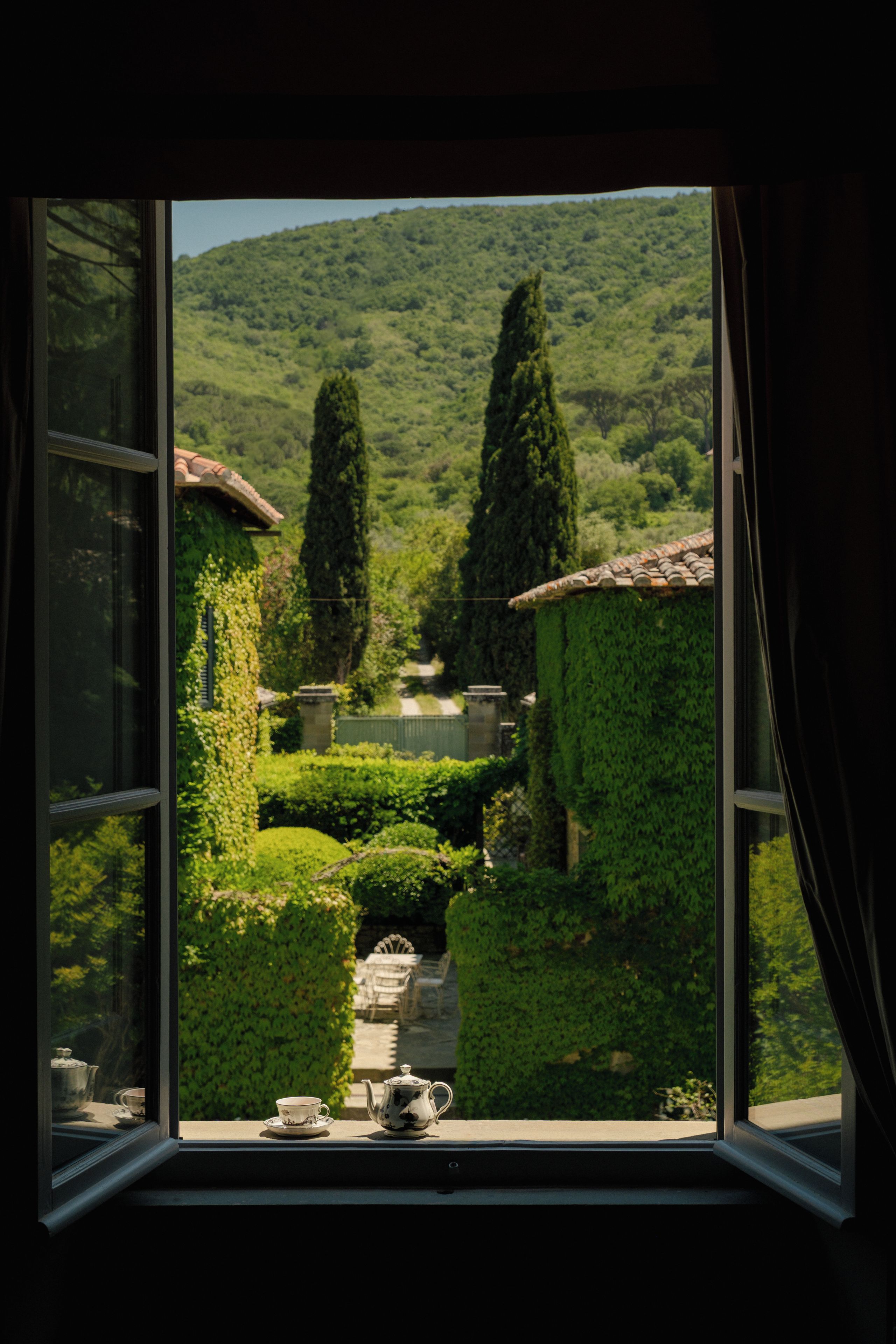 Vista desde una ventana a un jardín, que incluye un sendero, una mesa con tetera y sillas.