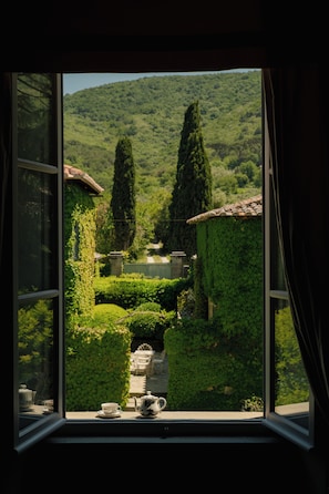 A view through a window with a garden, including a pathway, a table with a teapot, and chairs.