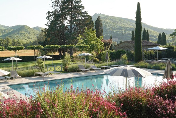 Una piscina con sillas de descanso y sombrillas, rodeada de follaje y colinas.