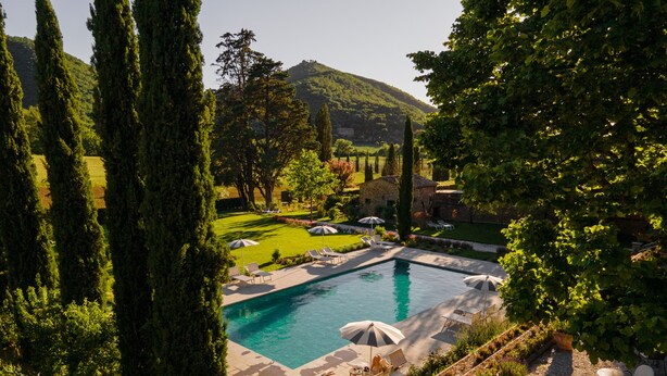 A swimming pool with lounge chairs and umbrellas, surrounded by greenery and a stone building in the background.