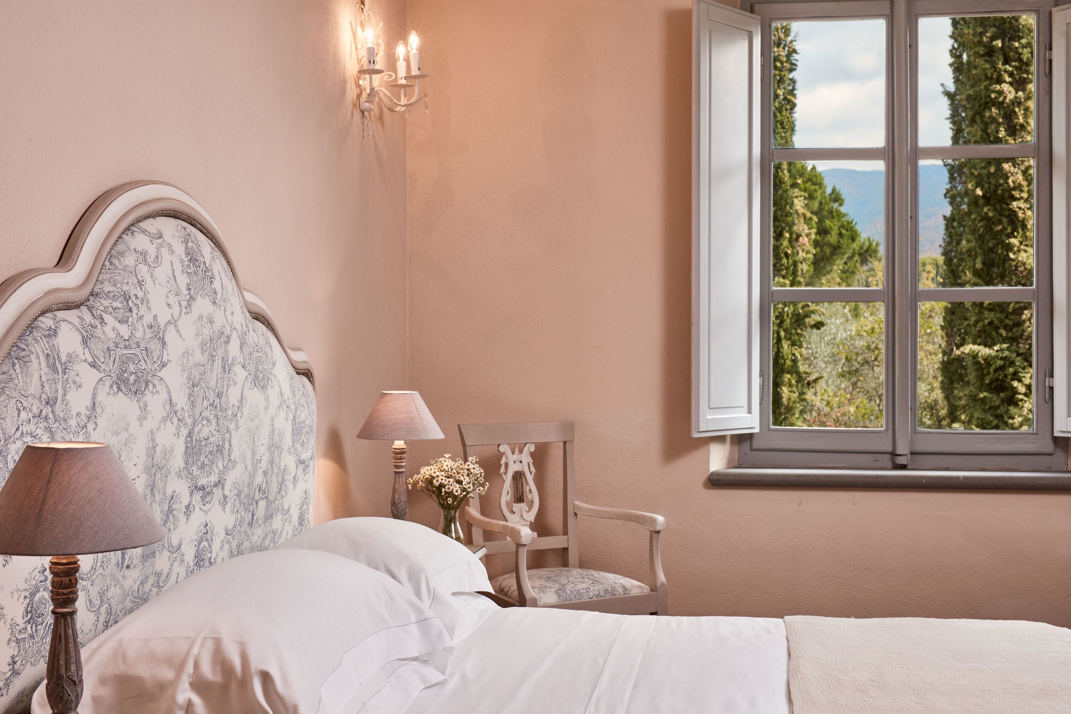 Une chambre à coucher comprenant un lit, une chaise, une lampe, un vase avec des fleurs et une fenêtre donnant sur des arbres.