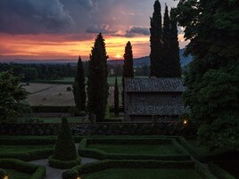 Un edificio de piedra rústico rodeado de setos cuidados y árboles altos, con un cielo al atardecer de fondo.