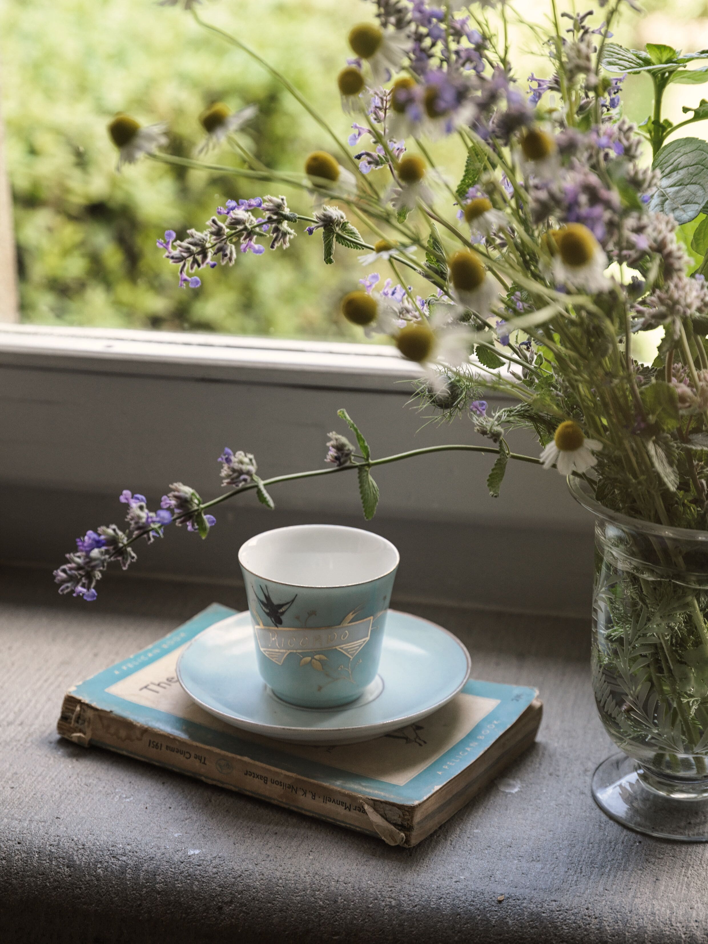 A window with a view of greenery, a blue cup with a saucer, and a book on a windowsill.