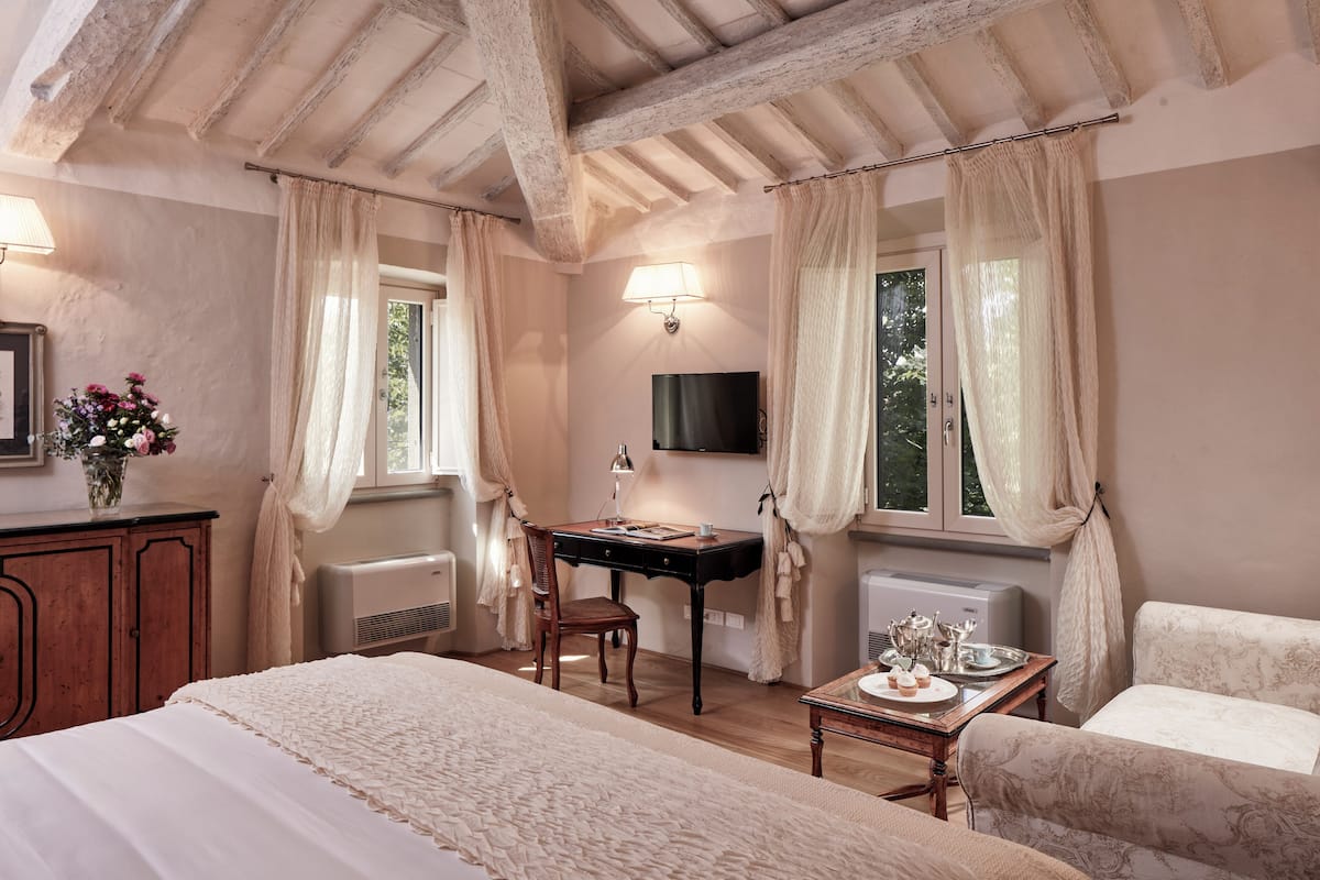 A bedroom with a bed, a desk, a chair, a table with a tray, a lamp, and a TV mounted on the wall.