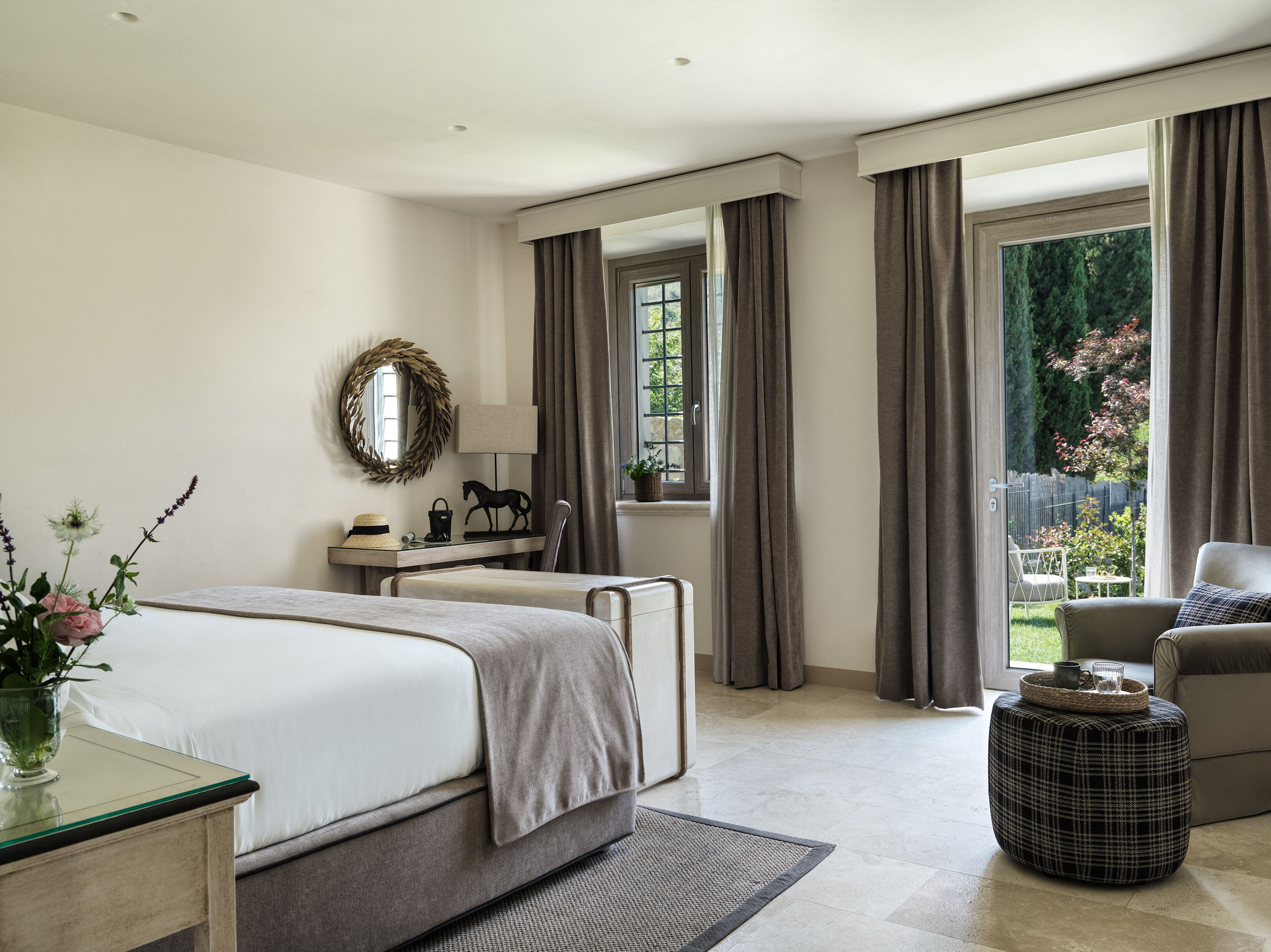 A modern bedroom with a large bed, a bedside table with a lamp, a mirror, and a view of a garden through a window.