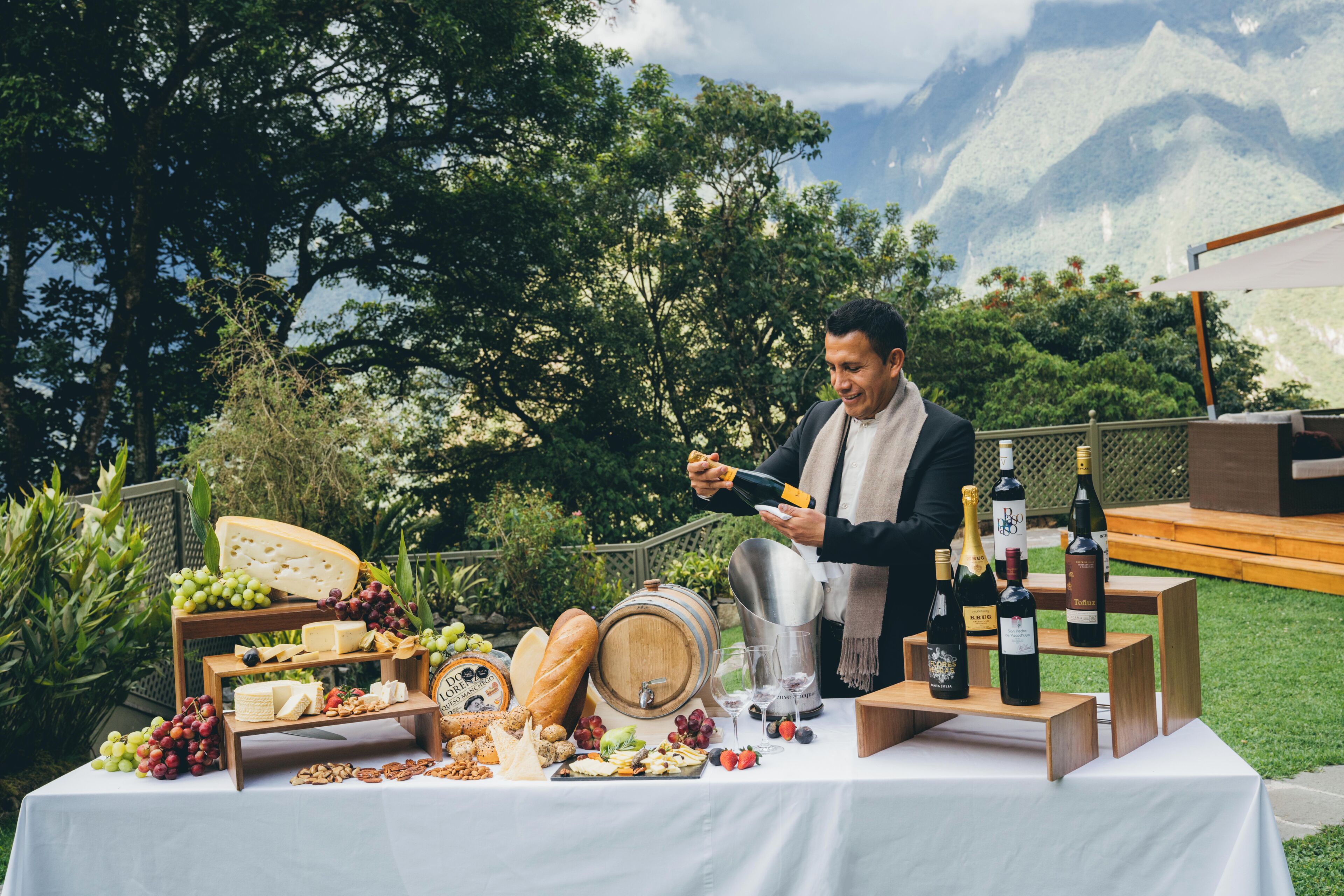 Un entorno al aire libre con una mesa dispuesta para una degustación de quesos y vinos, mostrando diferentes tipos de queso, uvas y botellas de vino.