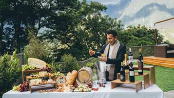 Un entorno al aire libre con una mesa dispuesta para una degustación de quesos y vinos, mostrando diferentes tipos de queso, uvas y botellas de vino.