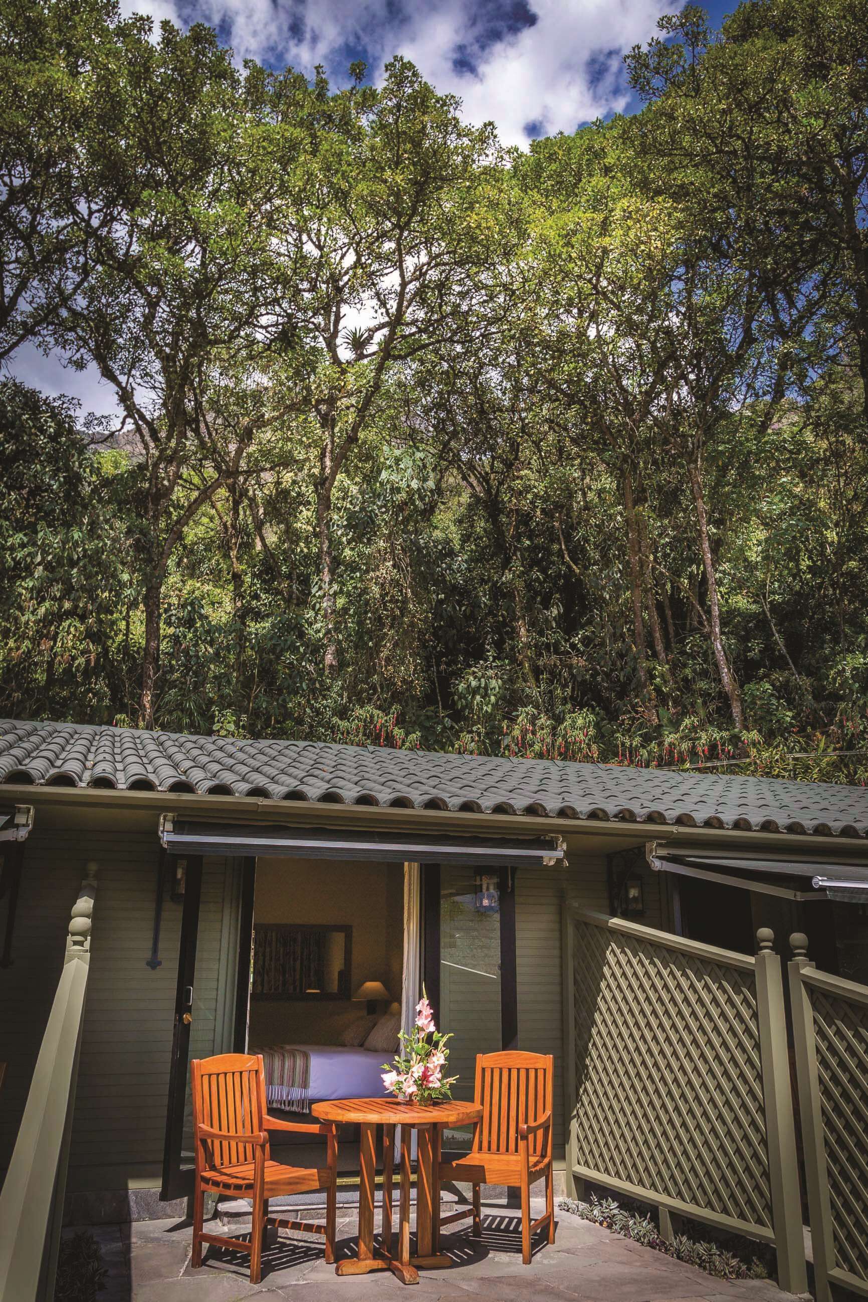 A wooden table and chairs outside a building with a tiled roof, surrounded by dense trees.