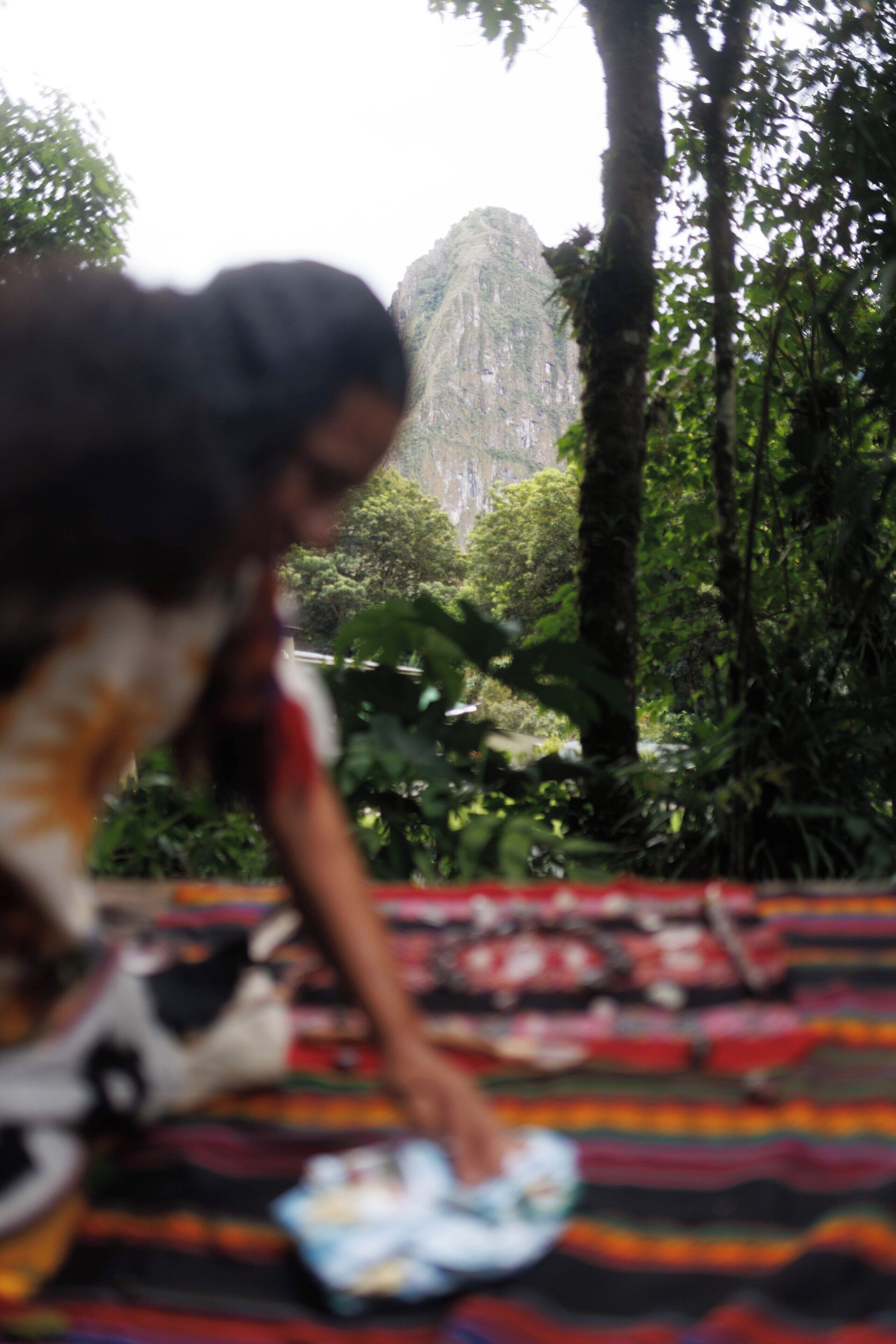 A person sitting on a colorful blanket outdoors, surrounded by lush greenery and a mountain in the background.