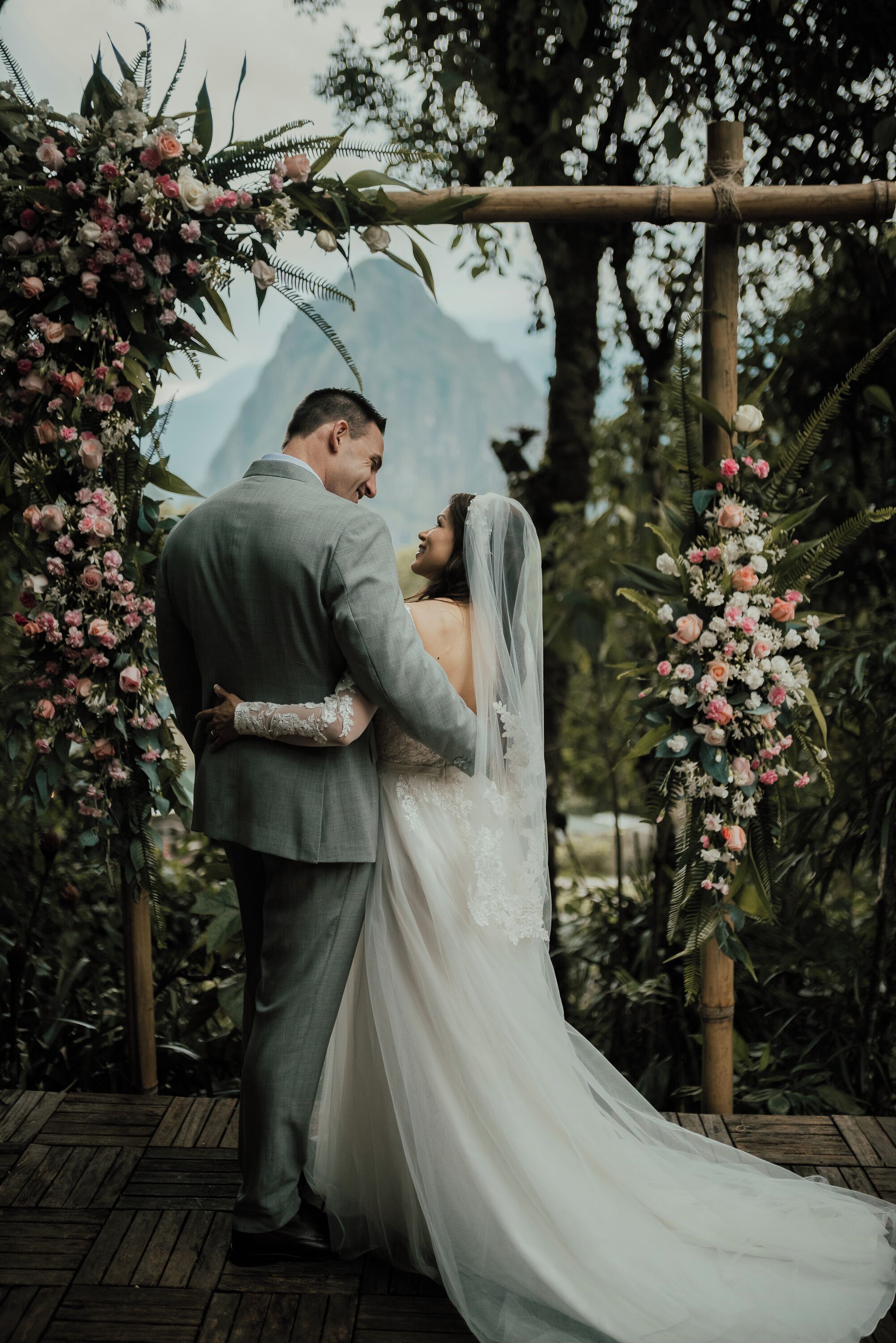 Una pareja abrazándose bajo un arco floral, con montañas al fondo.