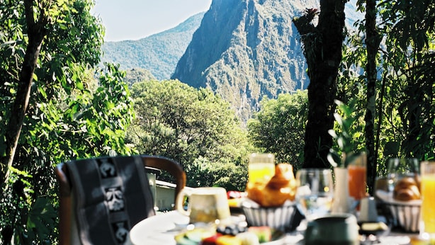 A table set outdoors with breakfast items, surrounded by lush greenery and a mountain in the background.