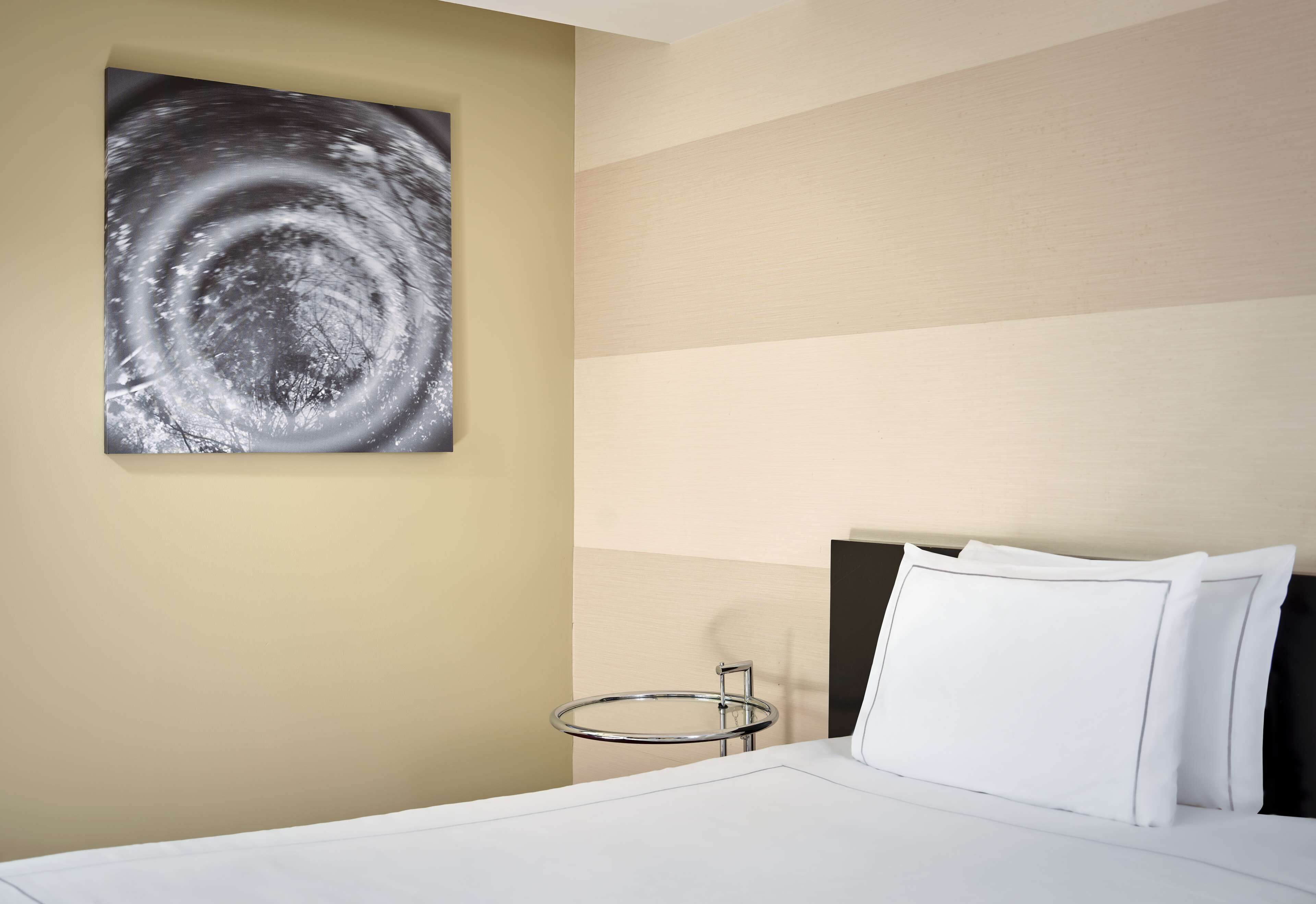 A hotel room with a bed, a round bedside table, a wall-mounted artwork, and a nightstand.