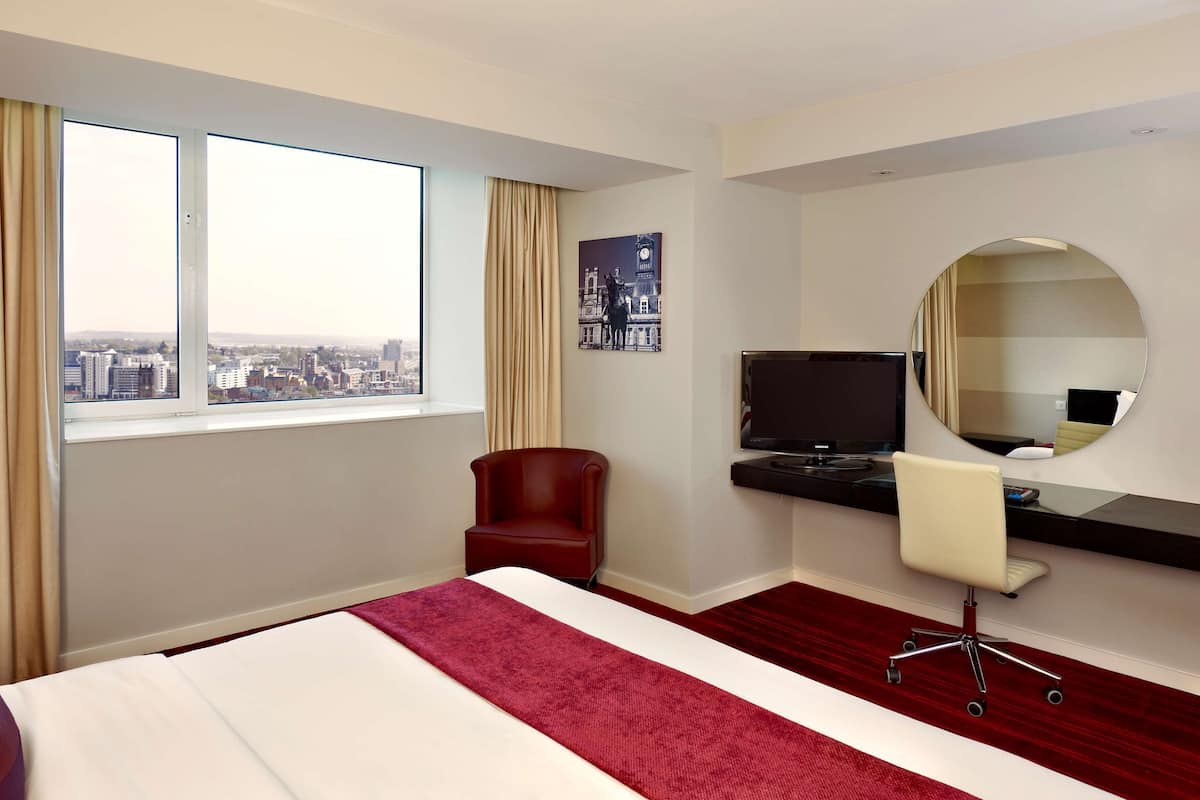 A hotel room with a bed, a desk with a TV, a chair, a large window with curtains, and a city view.
