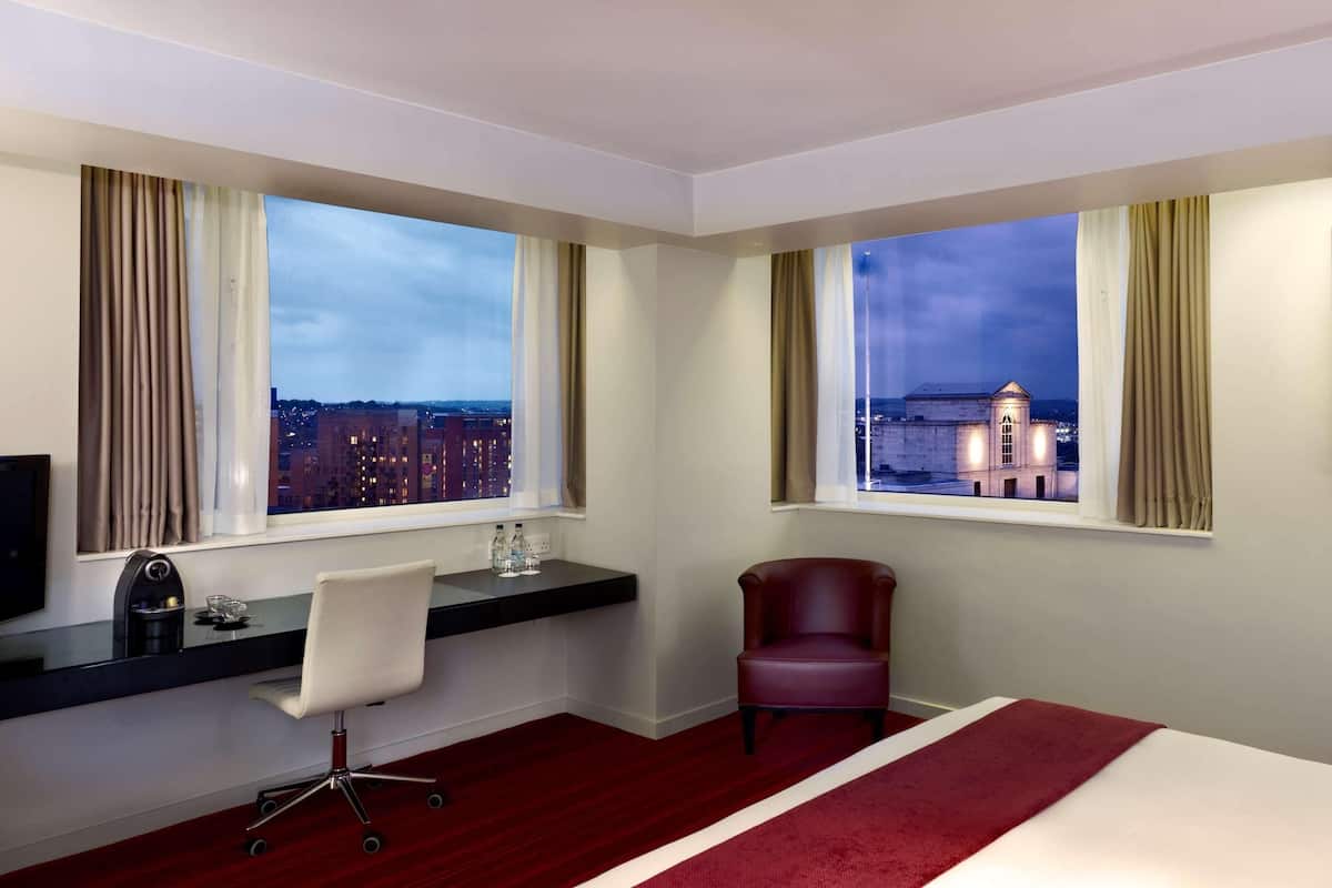 A hotel room with a bed, desk, chair, and two windows with city views.