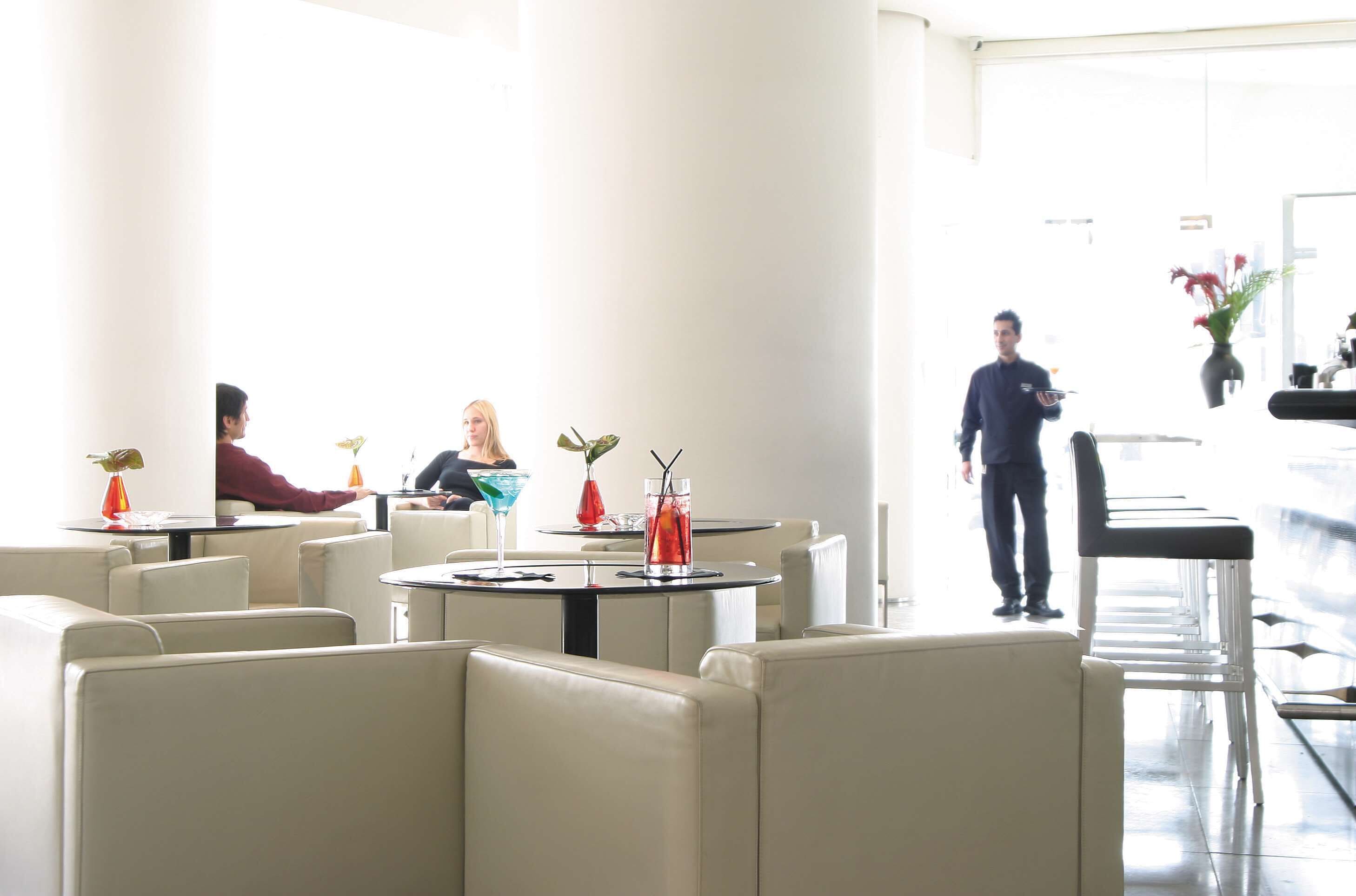 A modern restaurant with round tables and white chairs, a waiter in uniform, and a bright interior.