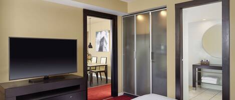 A hotel room with a flat-screen TV on a dark wooden dresser, a bed with white linens, a red carpet, a sliding glass door closet, and a bathroom with a mirror and sink.