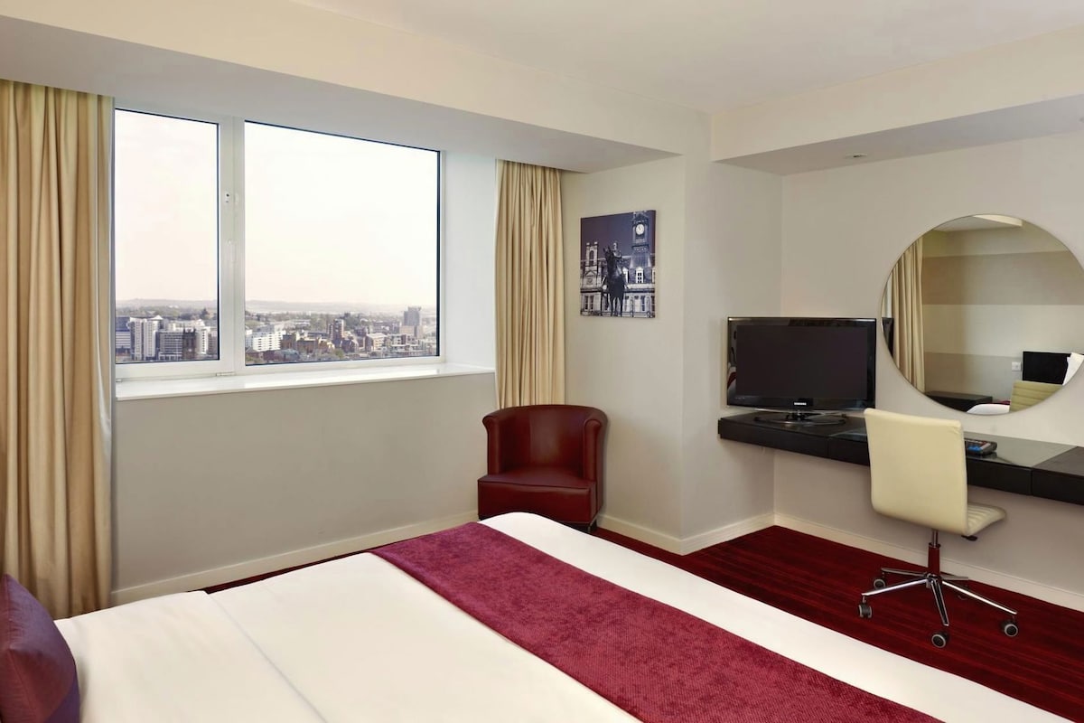 A hotel room with a bed, a desk with a computer, a chair, a large window with curtains, and a city view.