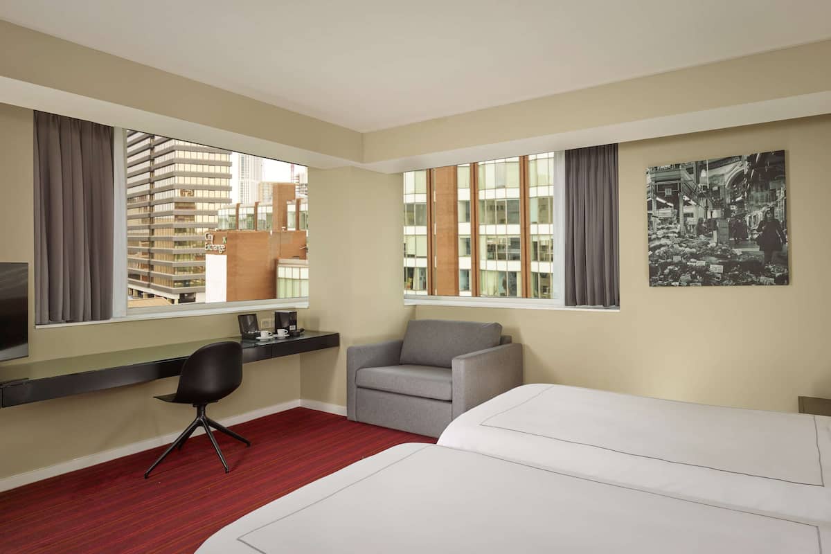 A hotel room with a bed, a desk with a chair, a coffee machine, and a large window with a city view.