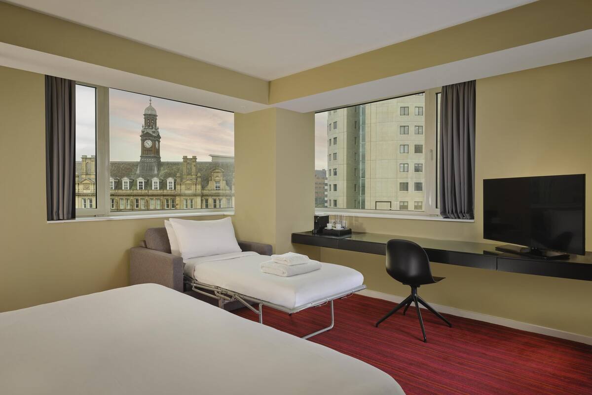 A hotel room with a bed, a desk, a chair, a TV, and large windows with a city view.