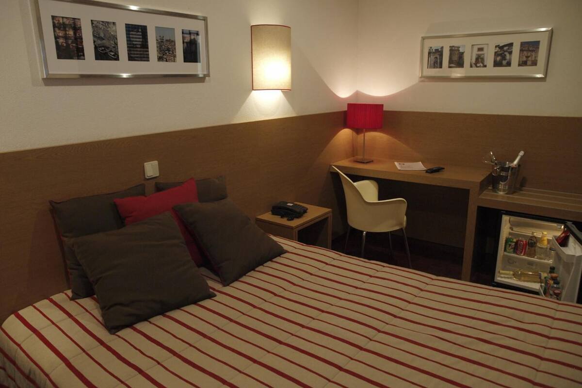A hotel room with a bed, a desk, a chair, a lamp, and a minibar.