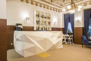 Uma área de bar com balcão geométrico branco, prateleiras com garrafas e um espaço de estar com cadeiras e um sofá.