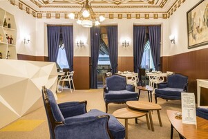 Um saguão de hotel com cadeiras estofadas em azul, um candelabro e uma área de bar com cadeiras e mesas brancas.