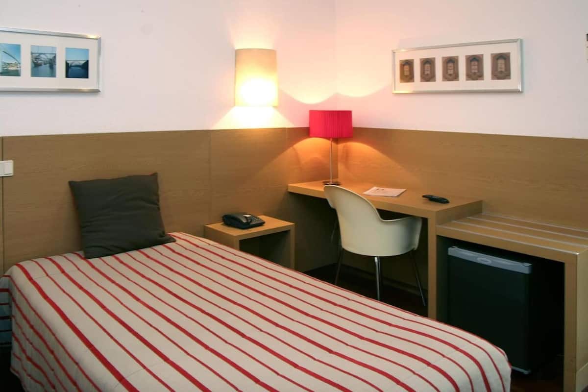 A hotel room with a bed, a desk, a chair, a lamp, a phone, and a small refrigerator.