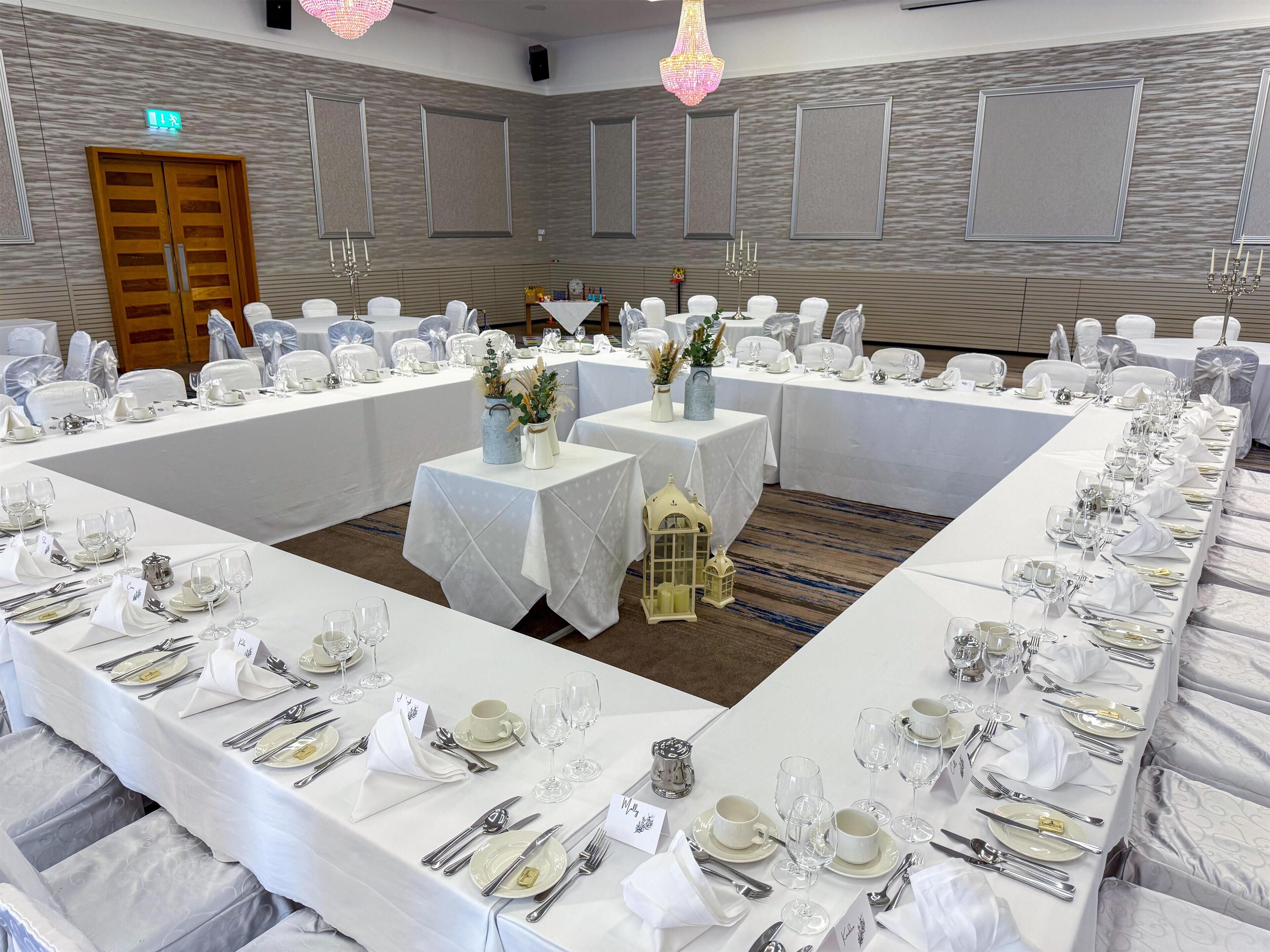 A banquet hall with long tables set for a formal event, featuring white tablecloths, silverware, and glassware.