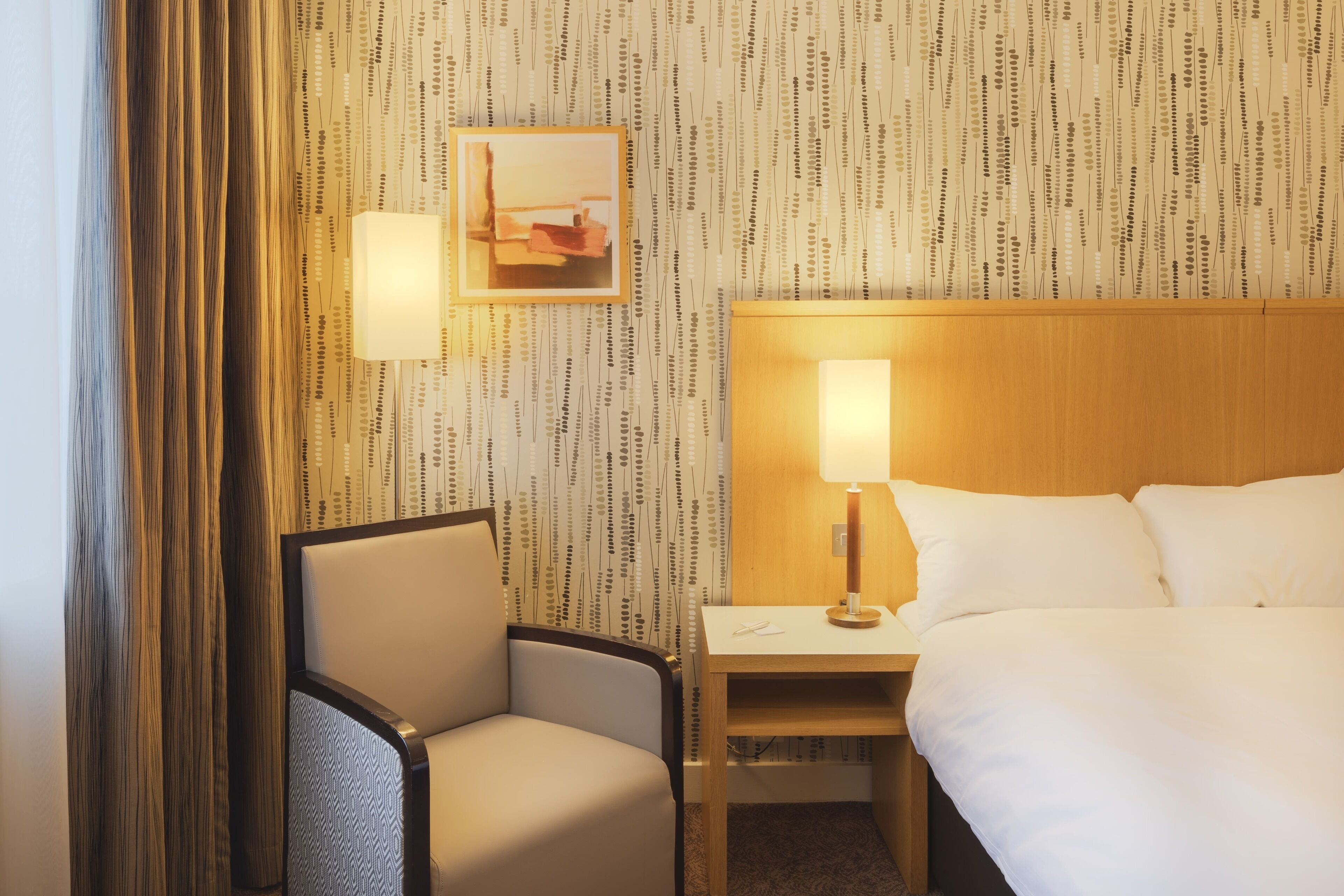 A hotel room with a bed, a chair, a nightstand, and a wall with a patterned wallpaper.