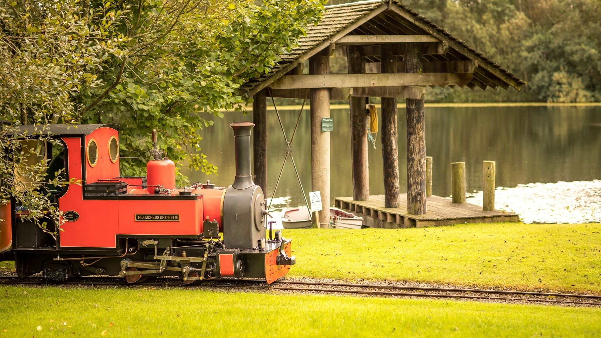 A red steam locomotive named "The Duchess of Daffin" on tracks near a wooden structure by a lake.
