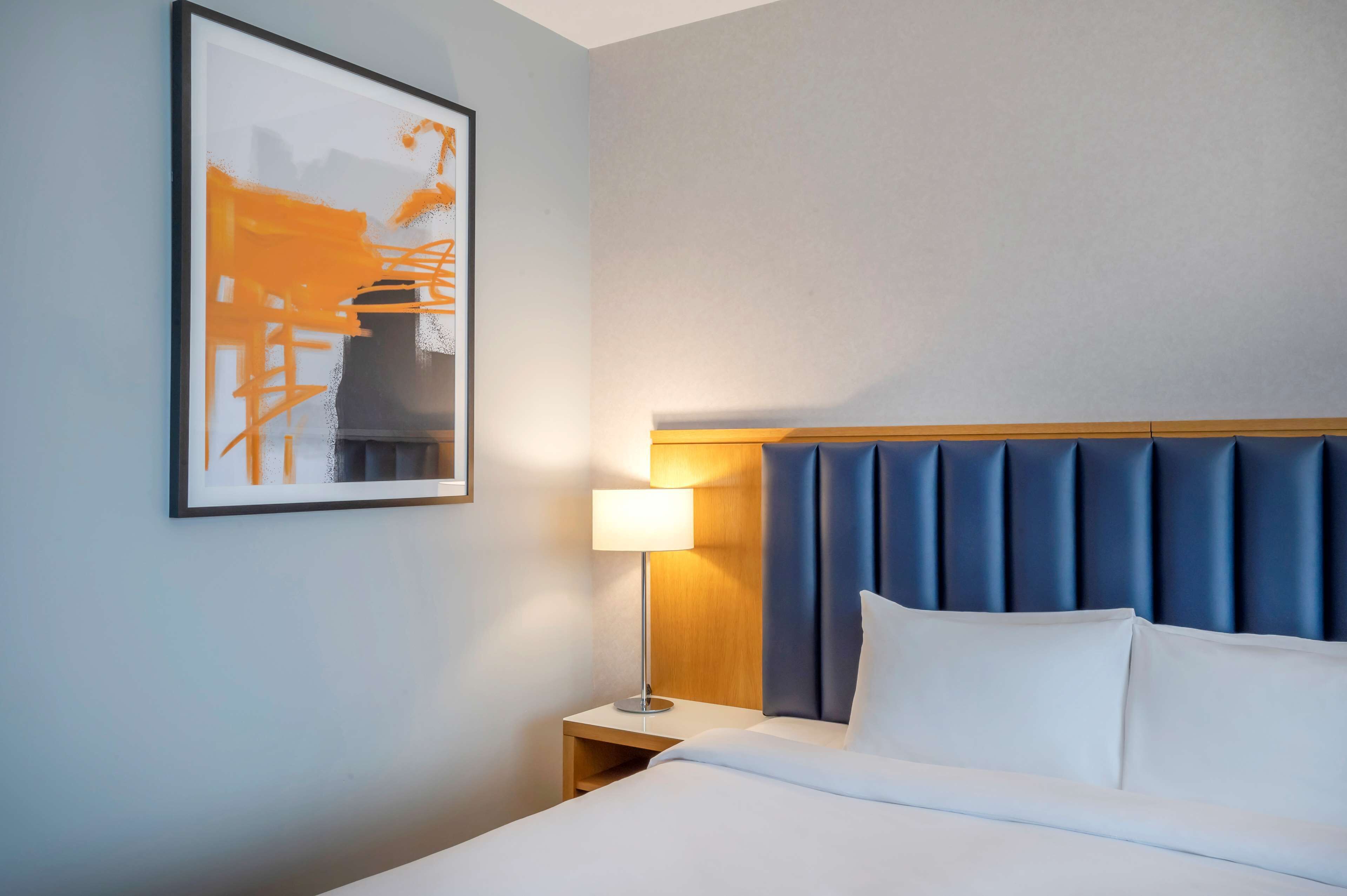 Et hotelværelse med seng, natbord, lampe, et billede i ramme på væggen og en blå sengegavl.