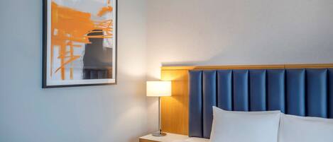 Et hotelværelse med seng, natbord, lampe, et billede i ramme på væggen og en blå sengegavl.