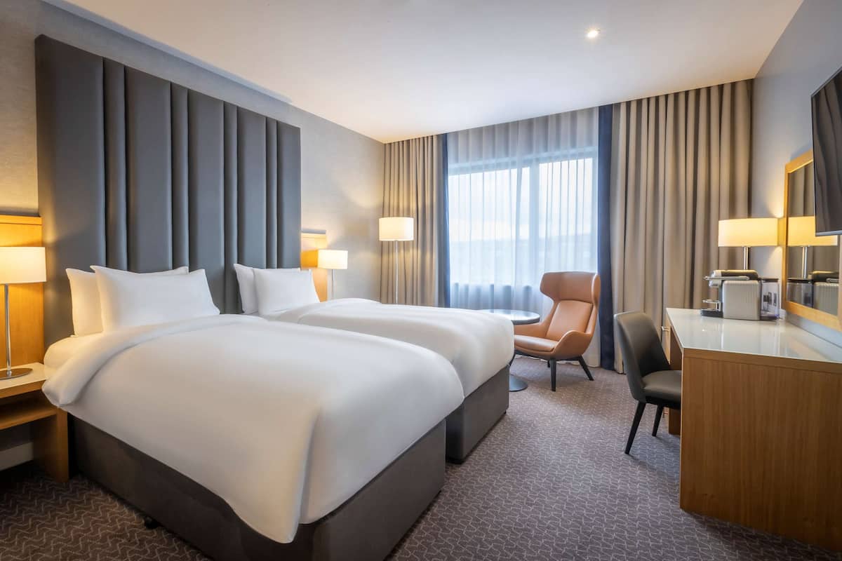 A hotel room with two beds, a desk, a chair, and a large window with curtains.
