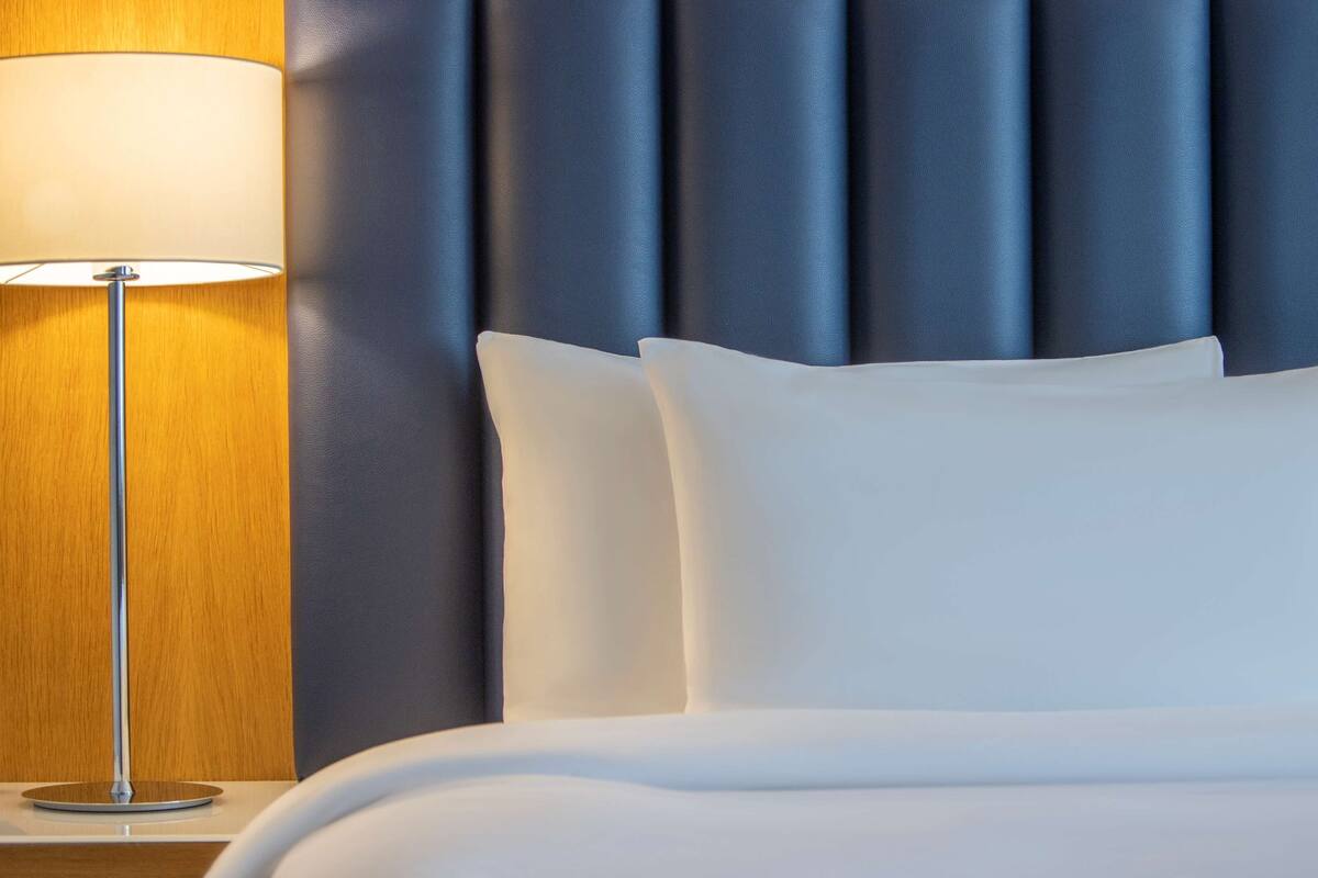 A neatly made bed with white linens and pillows, a modern lamp with a wooden base, and a blue upholstered headboard.