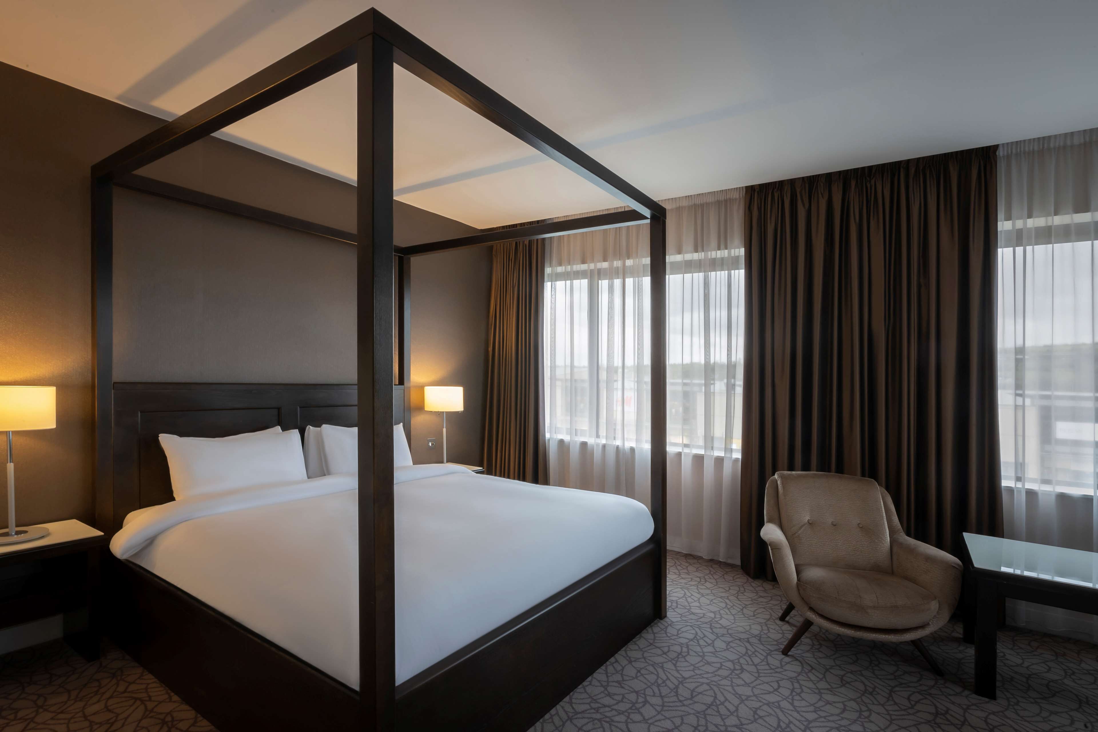 A four-poster bed with a dark headboard, two bedside lamps, a chair, and a table in a hotel room with large windows and curtains.