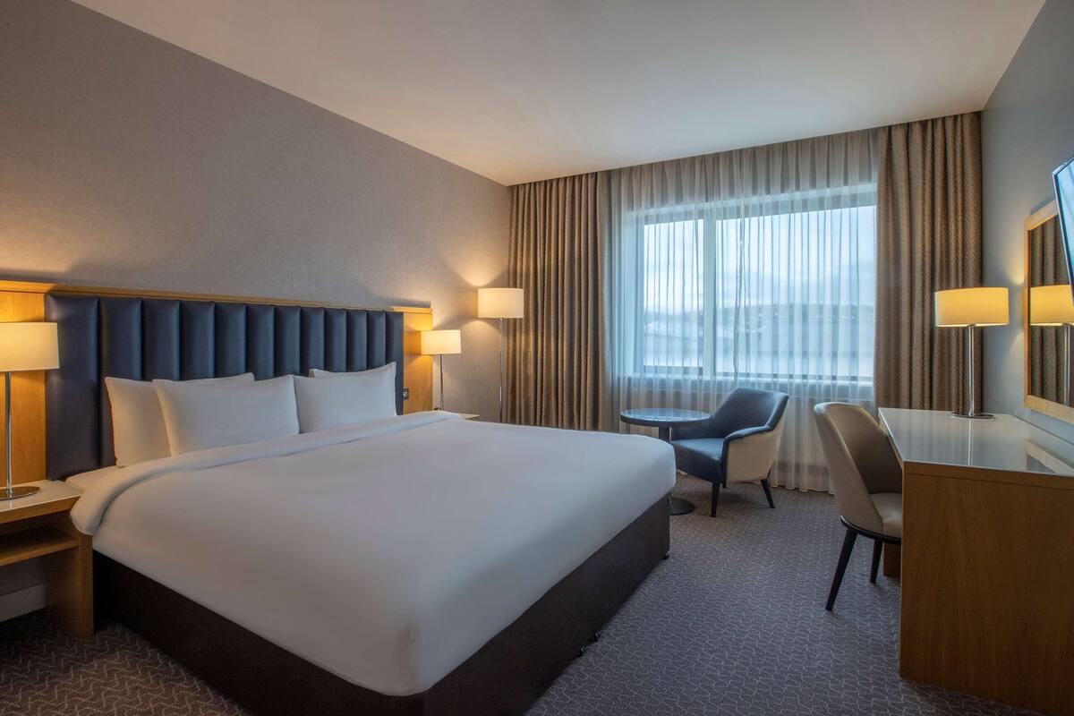 A hotel room with a large bed, two bedside tables with lamps, a desk with a chair, and a large window with curtains.