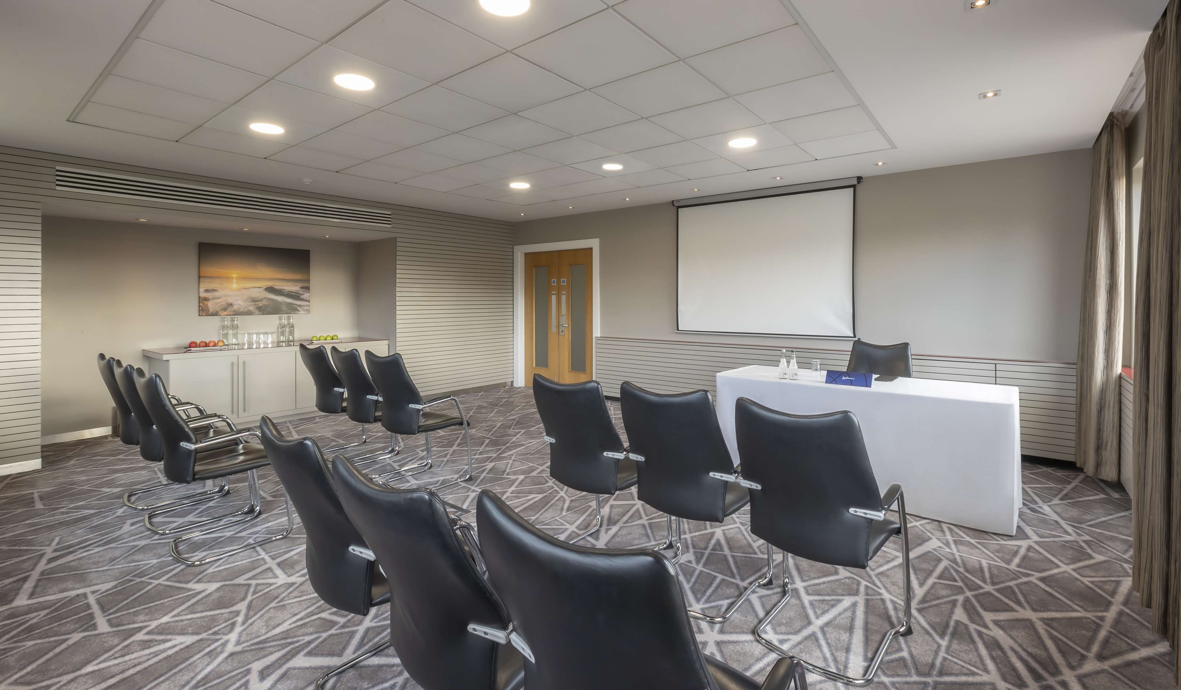Un salón de conferencias con una mesa blanca, sillas negras, una pantalla de proyector y un cuadro en la pared.