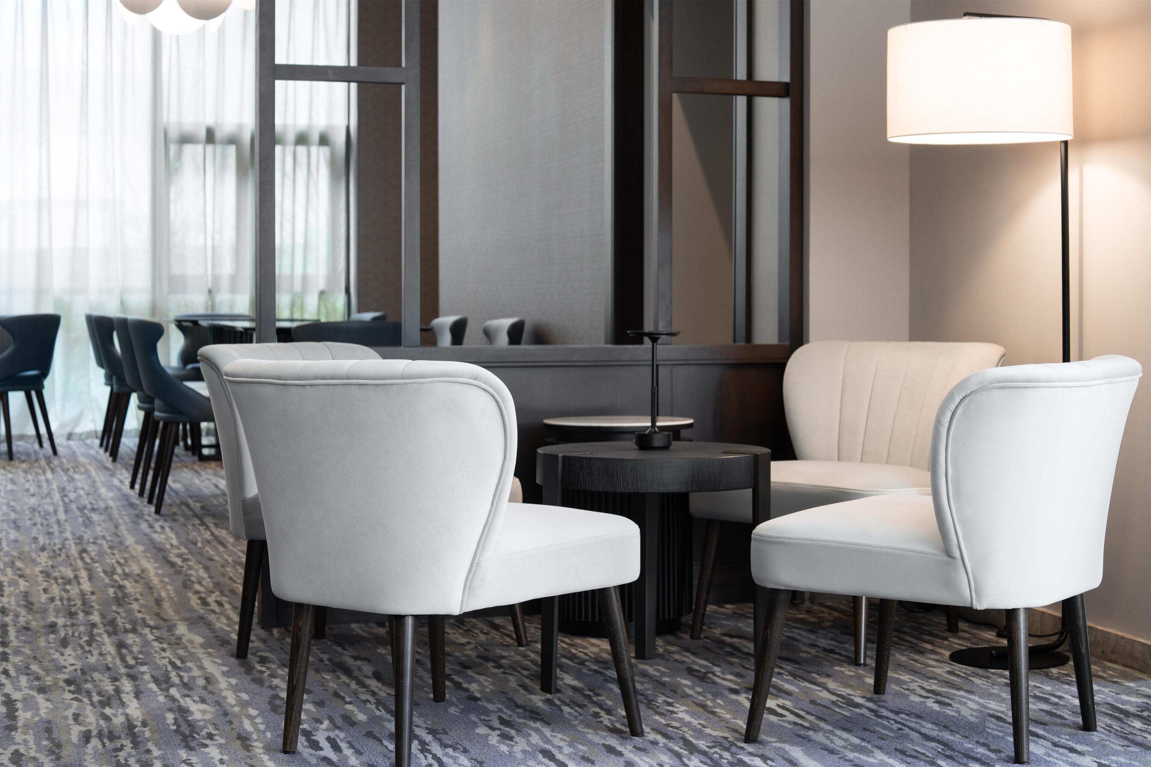 A modern hotel lobby with a patterned carpet, white leather chairs, a round table, and a floor lamp.