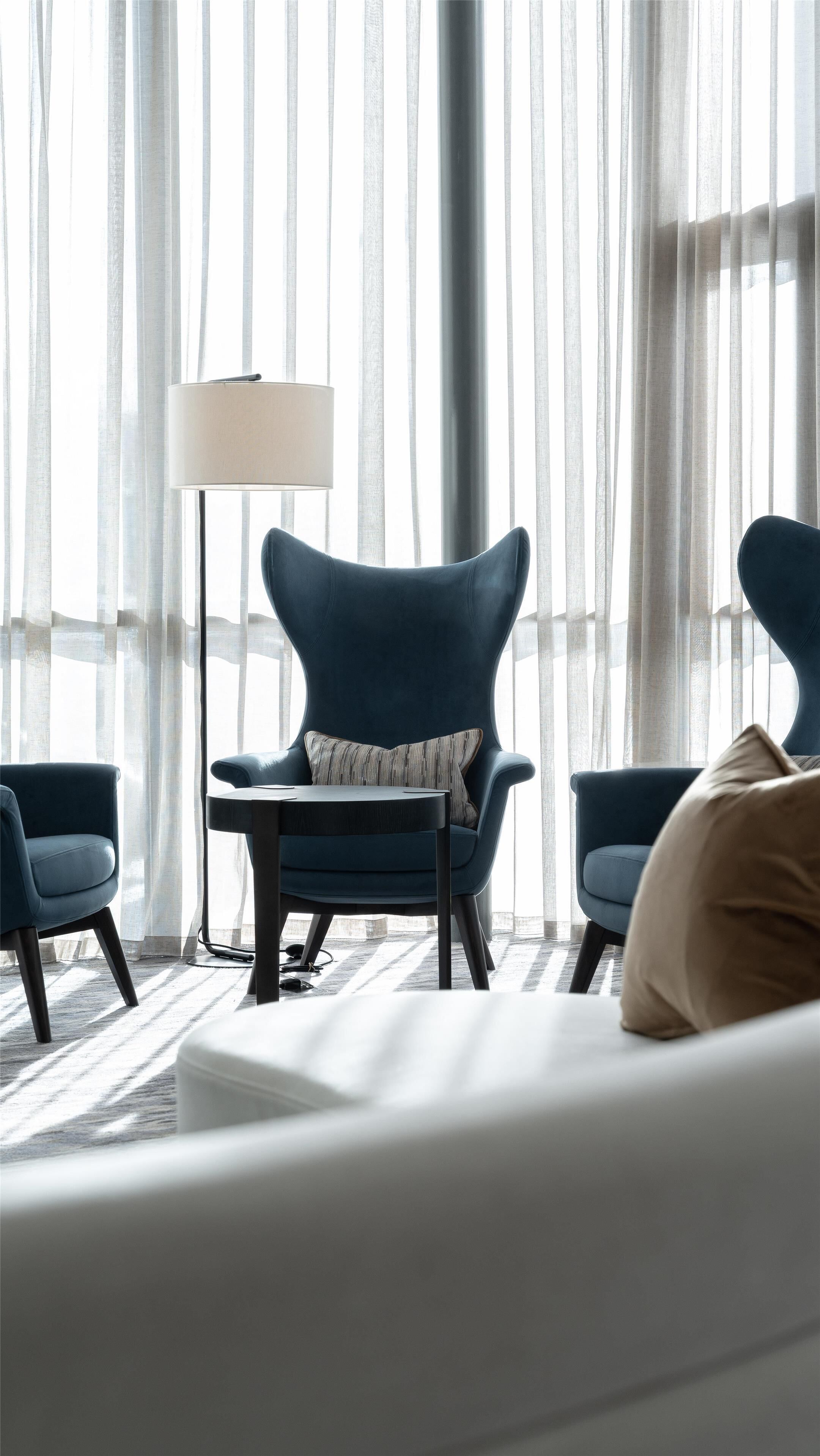 Una habitación de hotel moderna con un ventanal grande, una lámpara de pie y dos sillones azules.