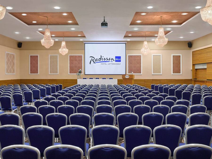 Un salón de conferencias con hileras de sillas azules, una pantalla grande mostrando
